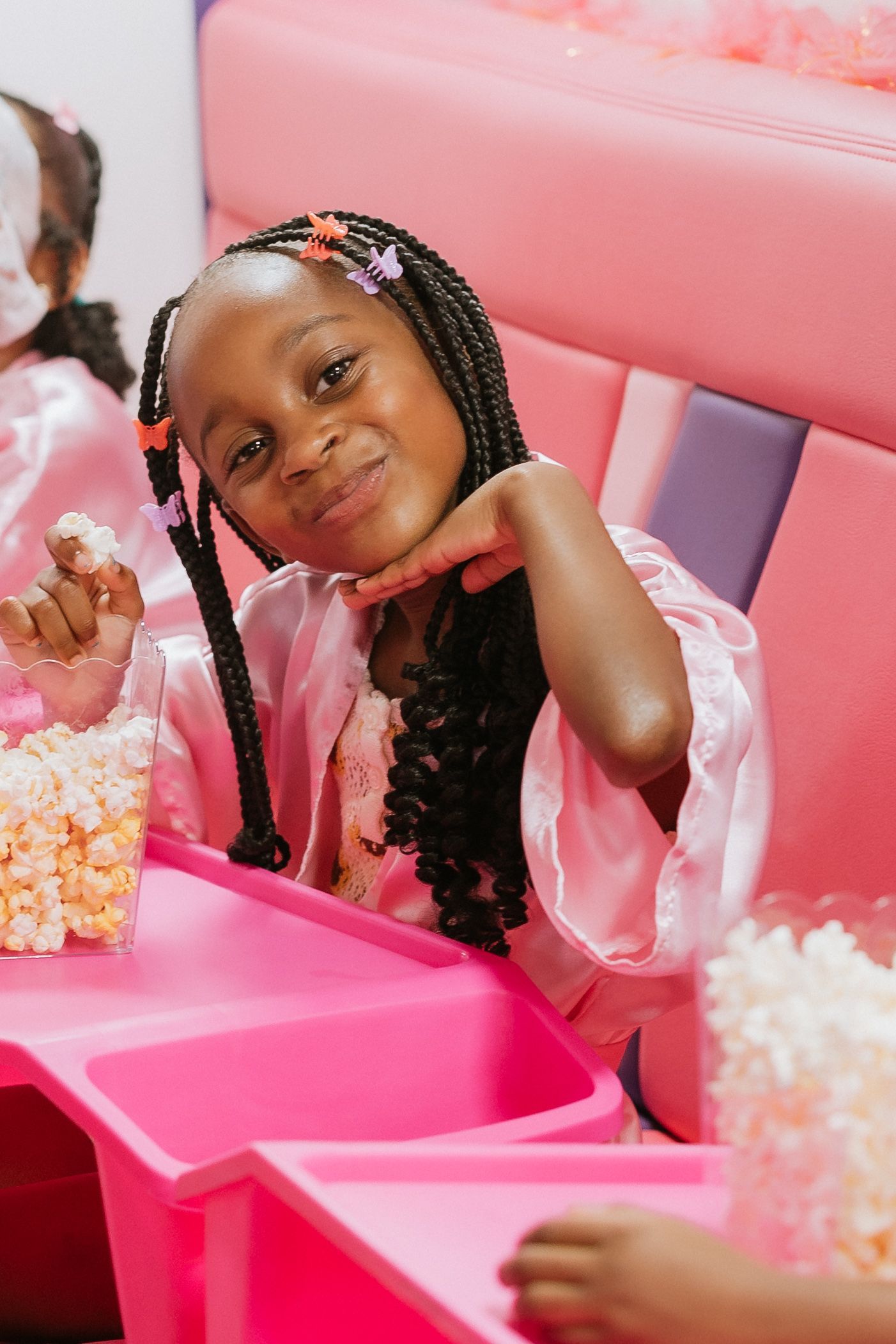 A little girl is sitting in a pink chair eating popcorn.