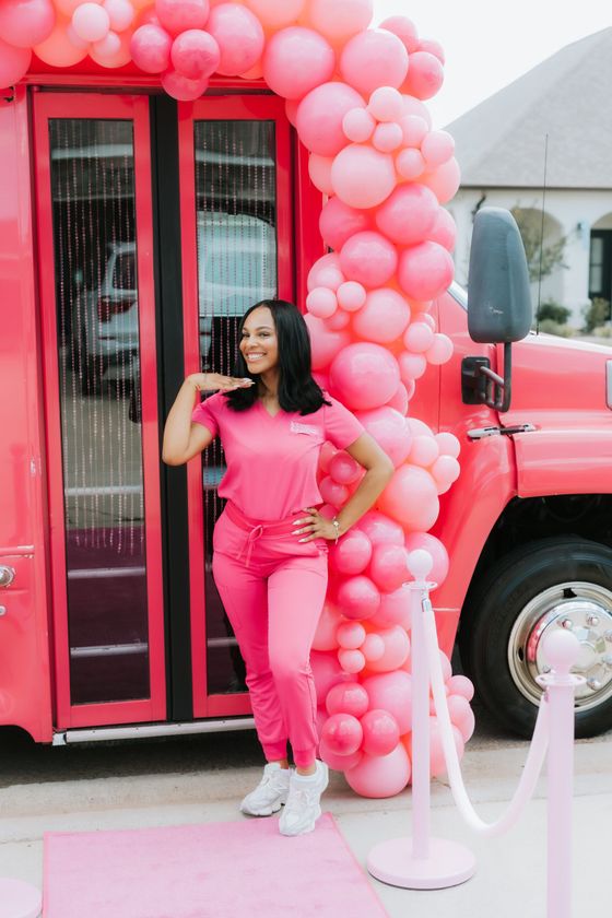 A woman in pink scrubs is standing in front of a pink bus decorated with pink balloons.