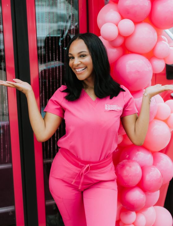 A woman in a pink scrub suit is standing in front of a pink balloon display.