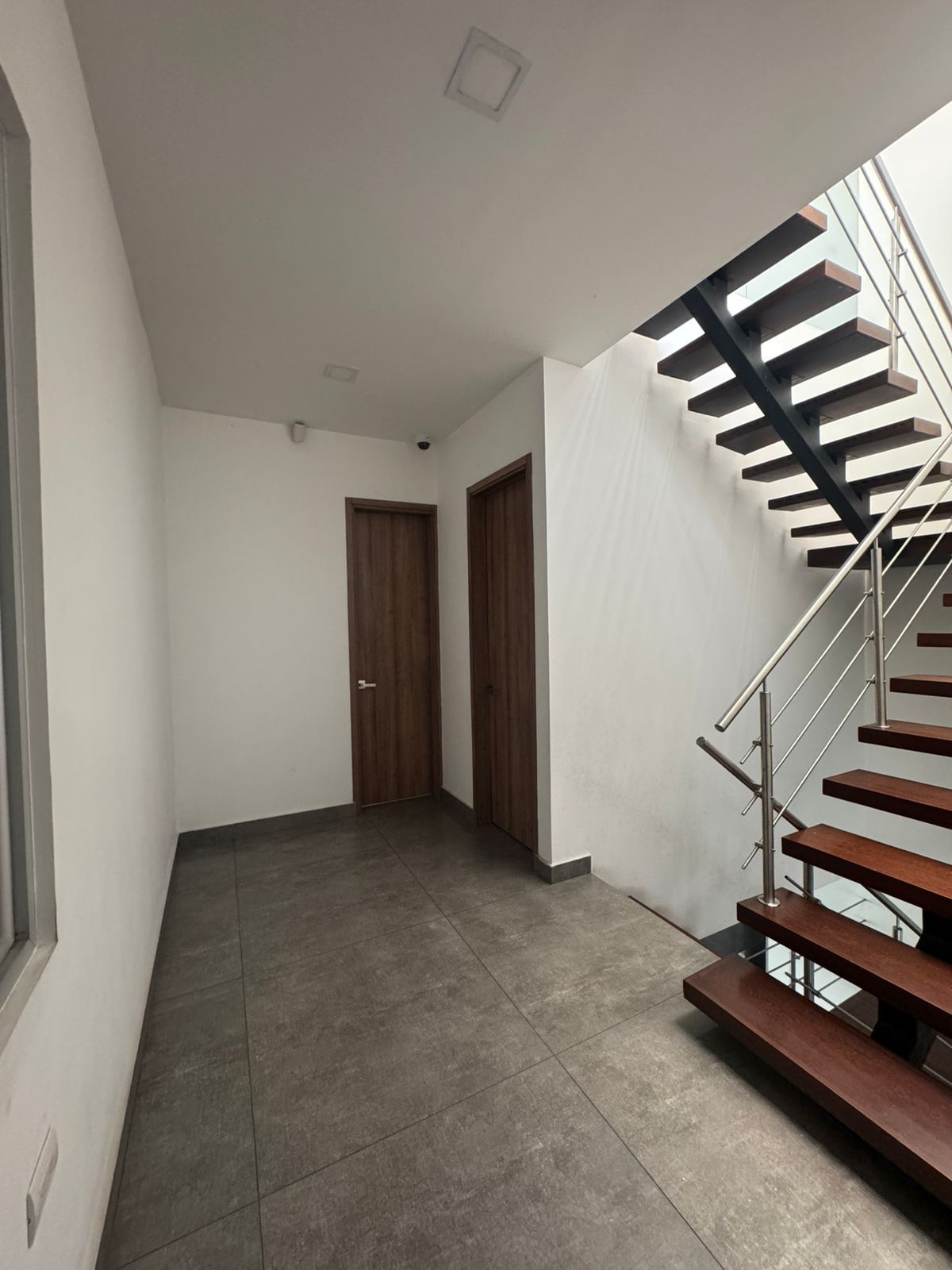 Pasillo con escaleras a la derecha y dos puertas cerradas. Escaleras de madera oscura, paredes claras y suelo de baldosas oscuras.