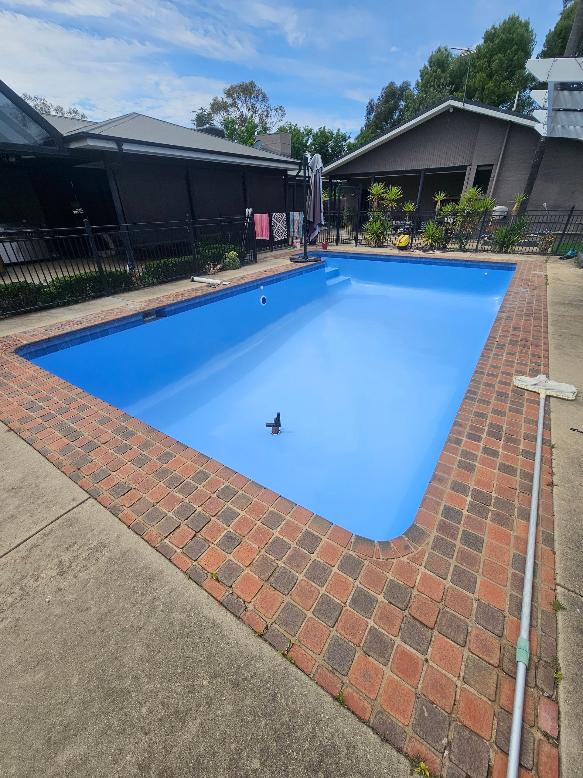 Rectangular blue swimming pool with brick border, surrounded by concrete. Buildings and trees in the background — Riverina Epoxy Flooring In Glenfield Park, NSW