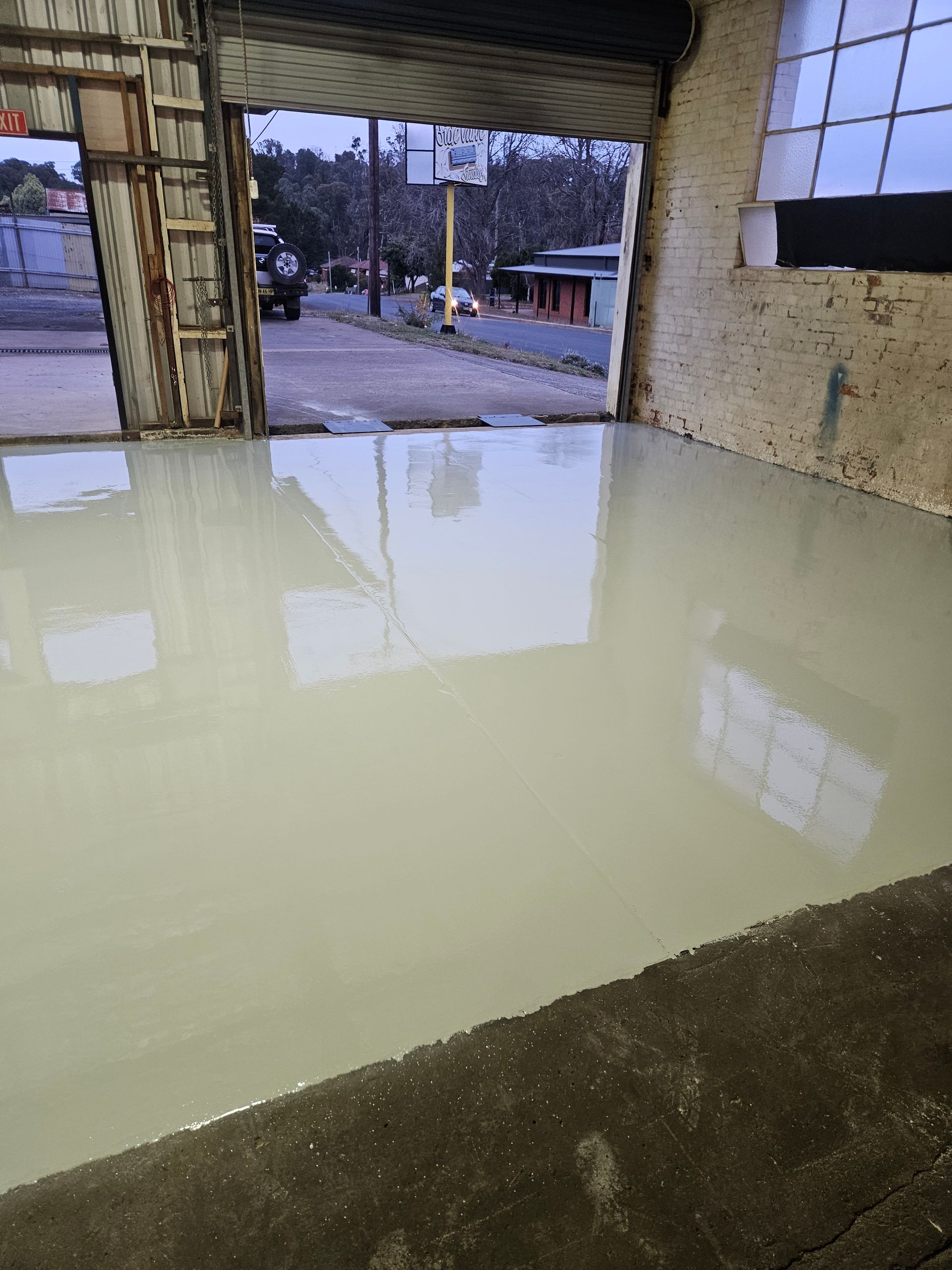 Indoor view of a concrete floor covered in a wet, pale gray coating, open doorway to street and buildings — Riverina Epoxy Flooring In Glenfield Park, NSW