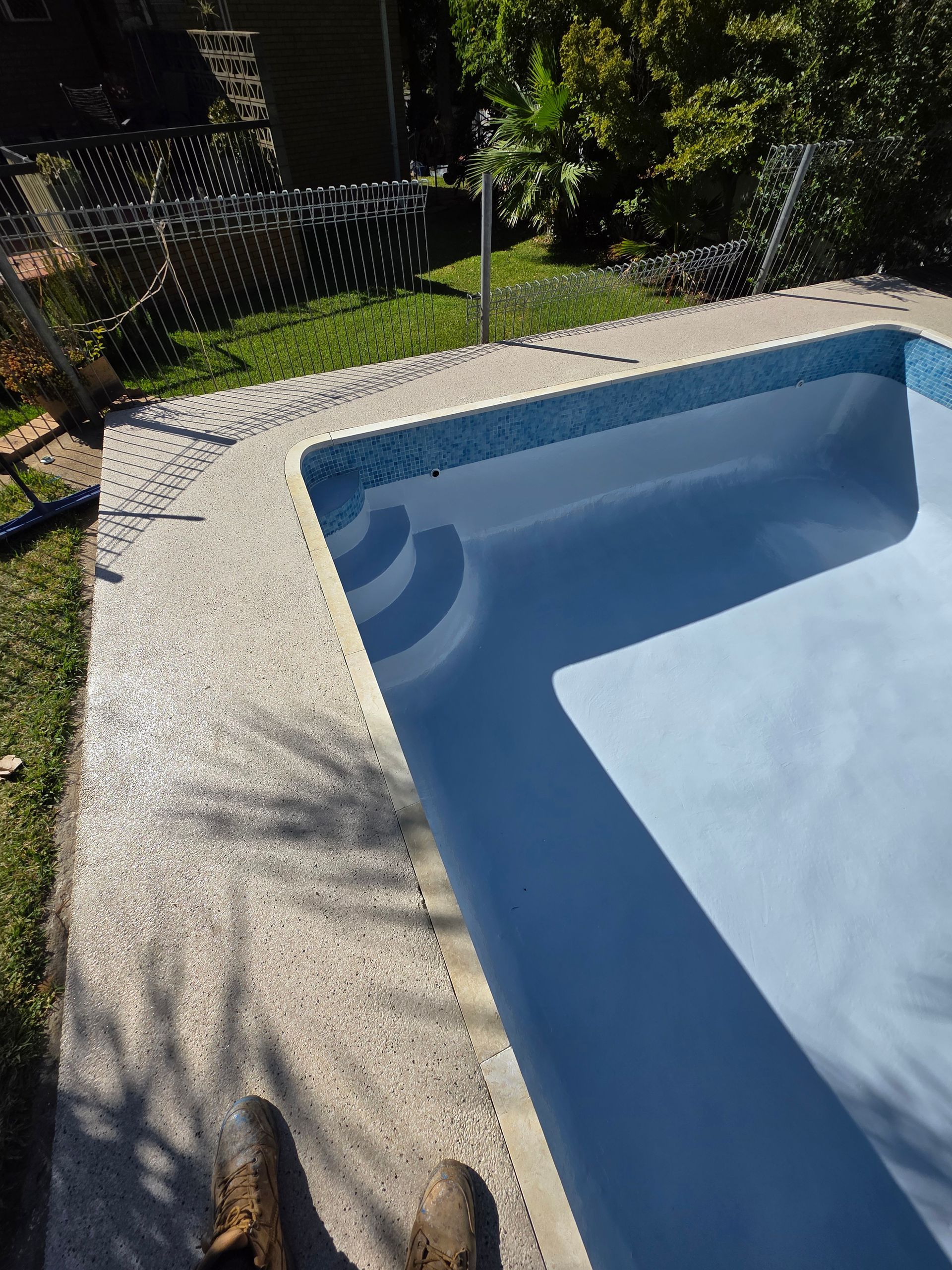 Concrete patio surrounds a blue-tiled swimming pool with steps, viewed from above. Green grass and fence in the background — Riverina Epoxy Flooring In Glenfield Park, NSW