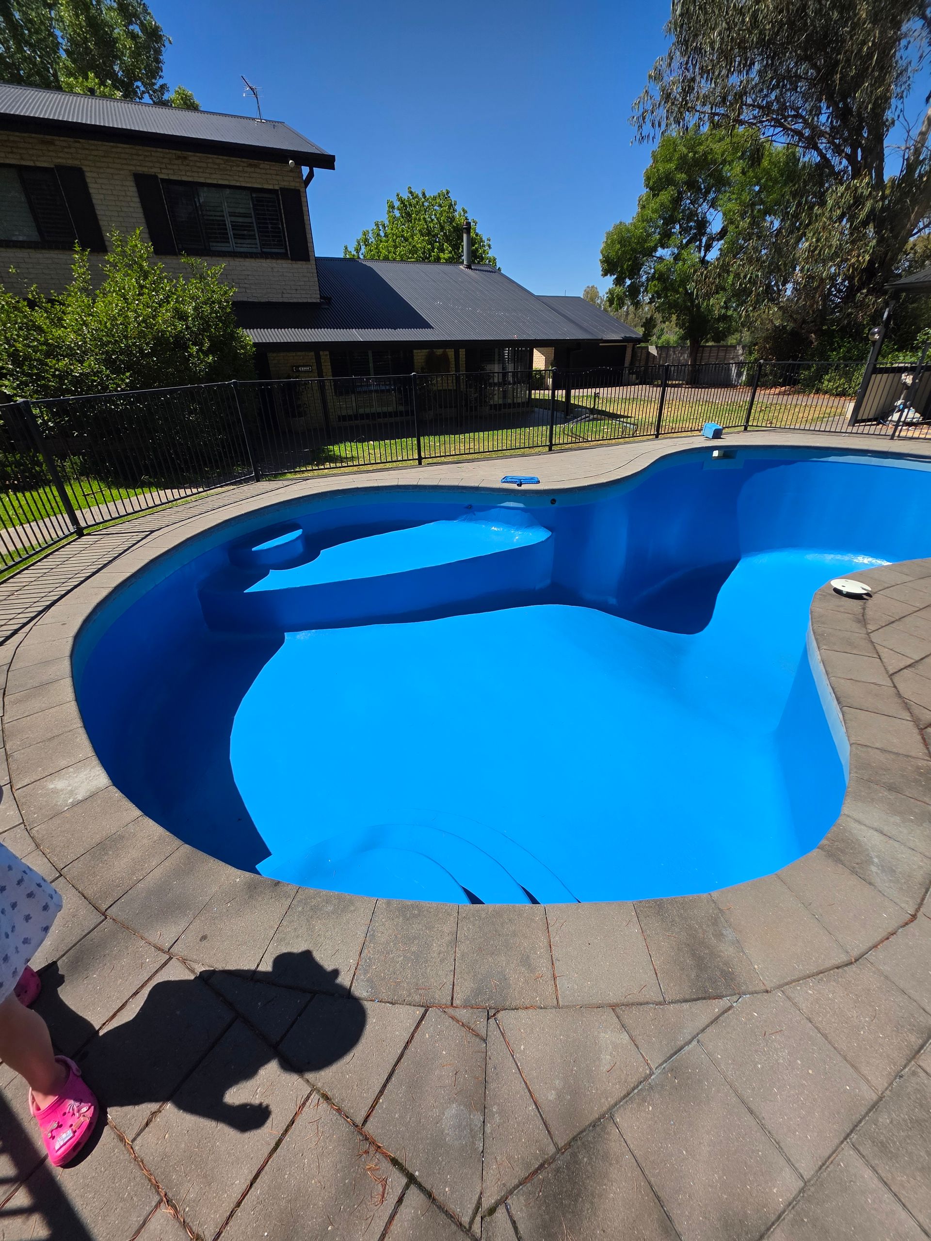 A bright blue, empty kidney-shaped swimming pool surrounded by brick and a fence, with a house in the background — Riverina Epoxy Flooring In Glenfield Park, NSW