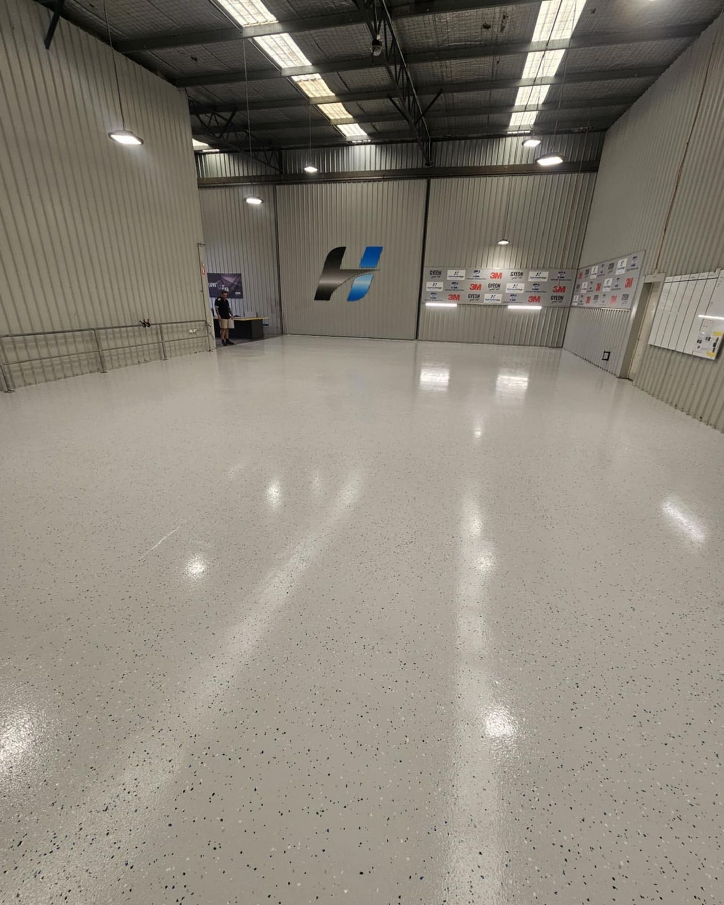 Empty warehouse with glossy white speckled floor, large logo on wall  — Riverina Epoxy Flooring In Glenfield Park, NSW