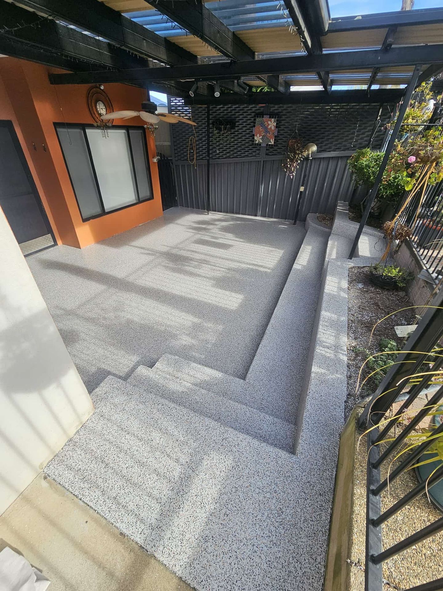 Concrete patio with steps, covered by a black pergola. Orange wall and black fence visible — Riverina Epoxy Flooring In Glenfield Park, NSW