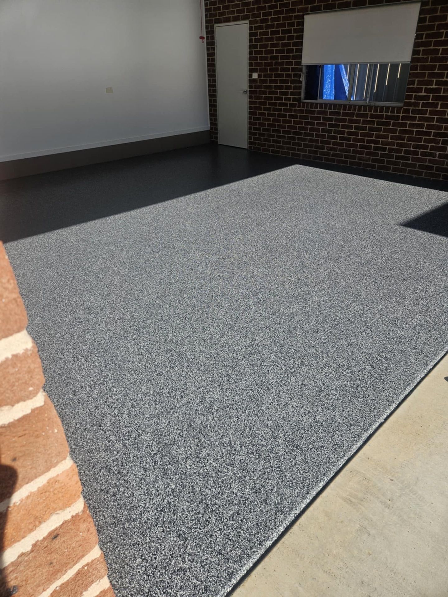 Garage with a speckled gray epoxy floor, brick walls, and a closed white door — Riverina Epoxy Flooring In Glenfield Park, NSW