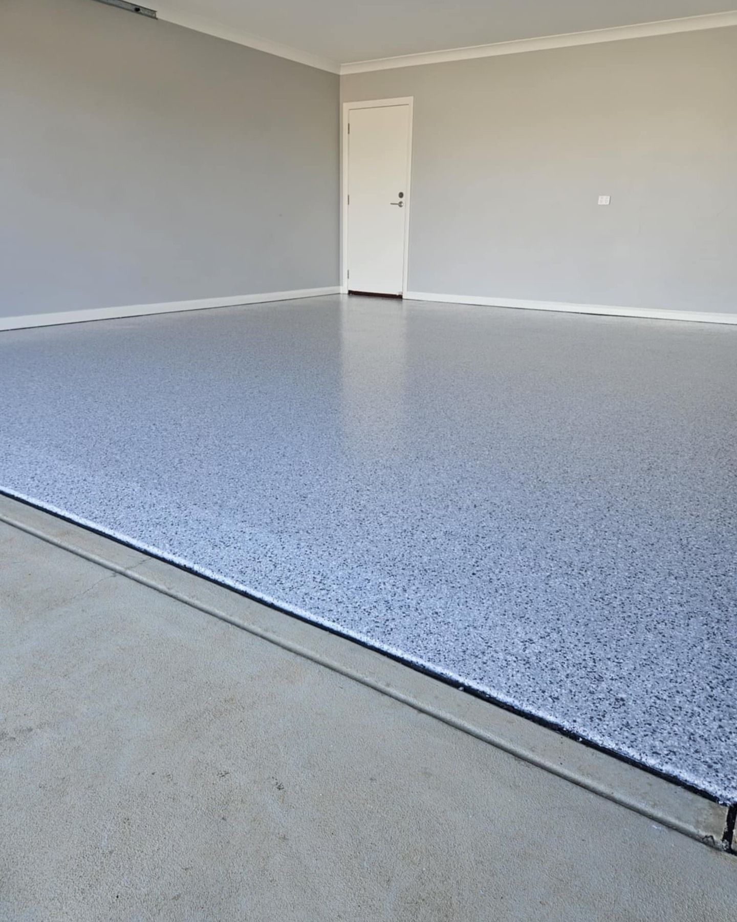 Garage with a speckled epoxy floor and grey walls. A white door is visible — Riverina Epoxy Flooring In Glenfield Park, NSW