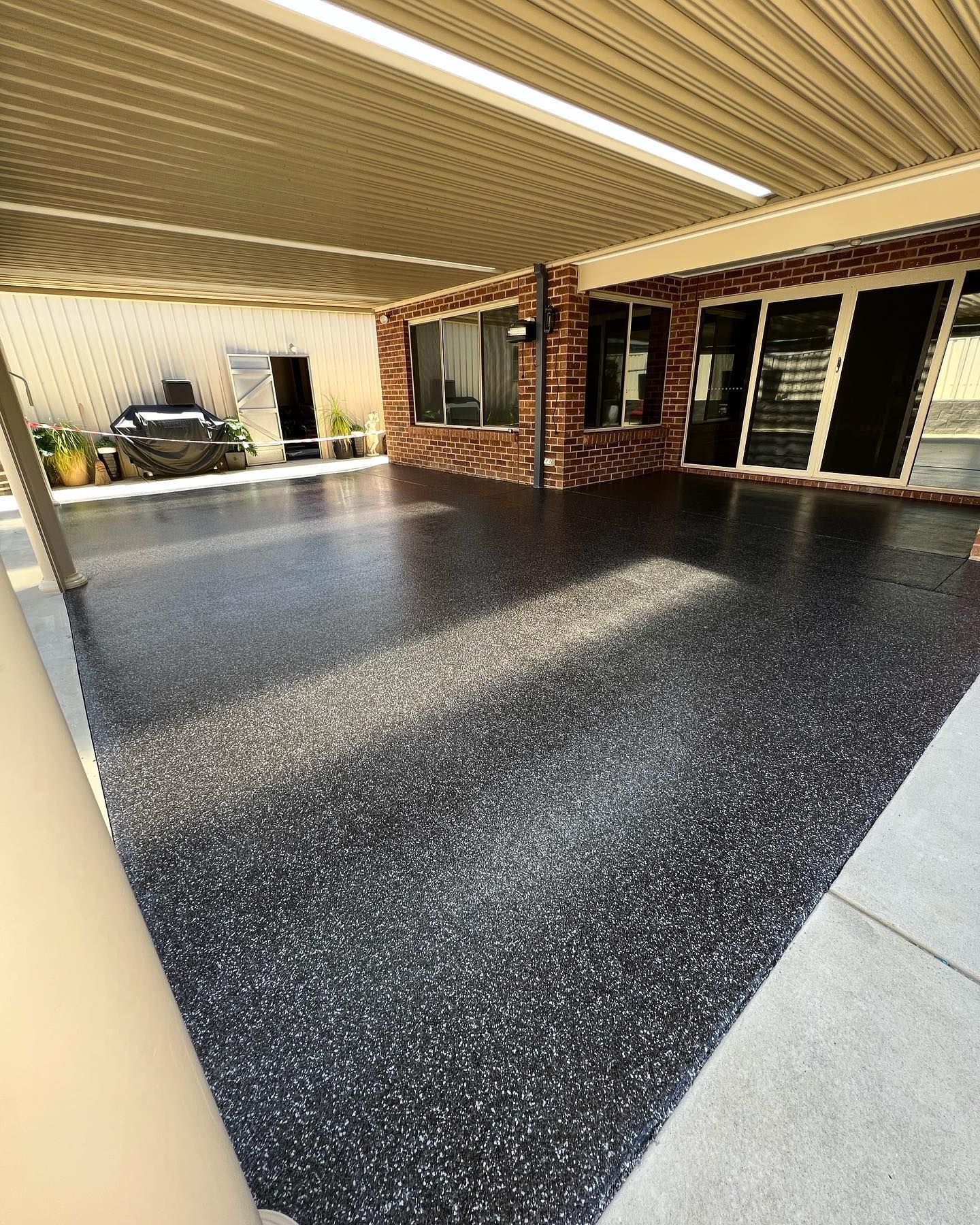 Covered patio with a glossy black and speckled floor, brick wall, and a garage door in the background — Riverina Epoxy Flooring In Glenfield Park, NSW