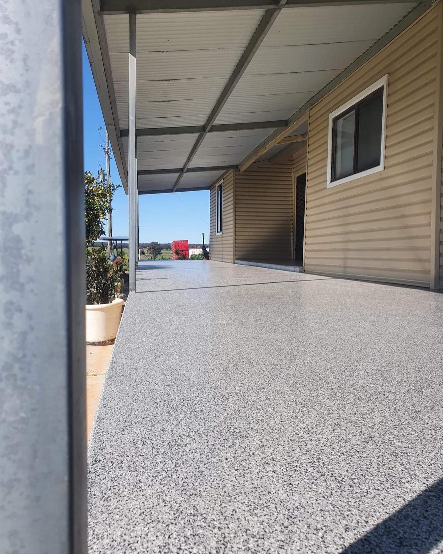 Covered concrete patio with gray speckled finish, tan siding, and a view of distant red object — Riverina Epoxy Flooring In Glenfield Park, NSW