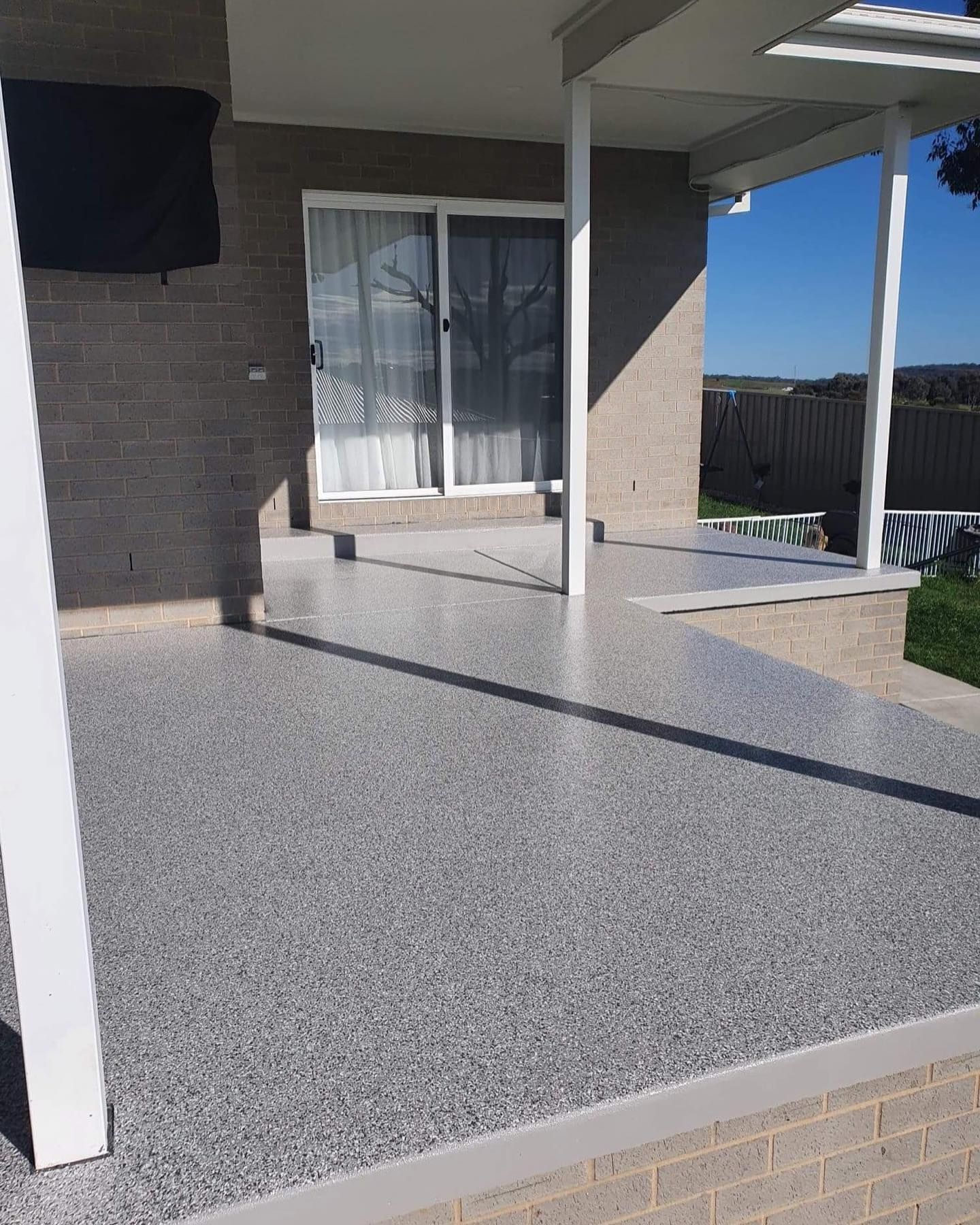 Gray speckled concrete patio with white pillars, next to a brick wall and sliding glass door — Riverina Epoxy Flooring In Glenfield Park, NSW