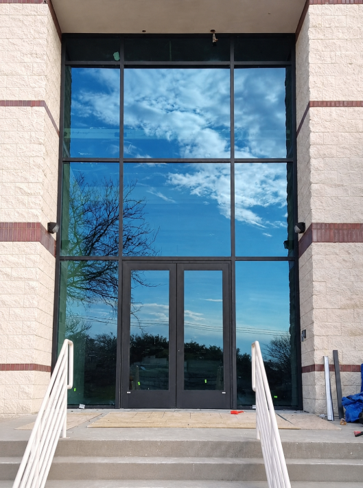Glass entrance doors reflecting a blue sky and clouds, framed by beige stone walls and steps.