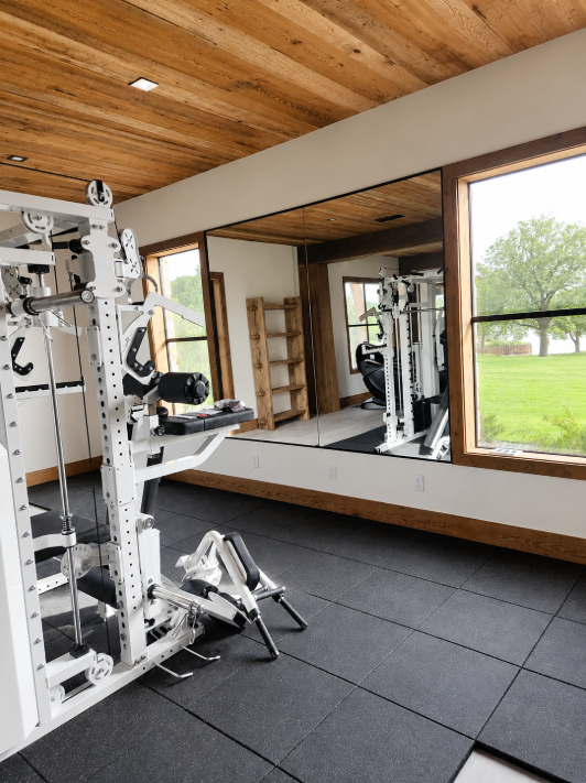 Modern home gym with weight machine, floor mats, wood ceiling, and large wall mirror overlooking a green lawn