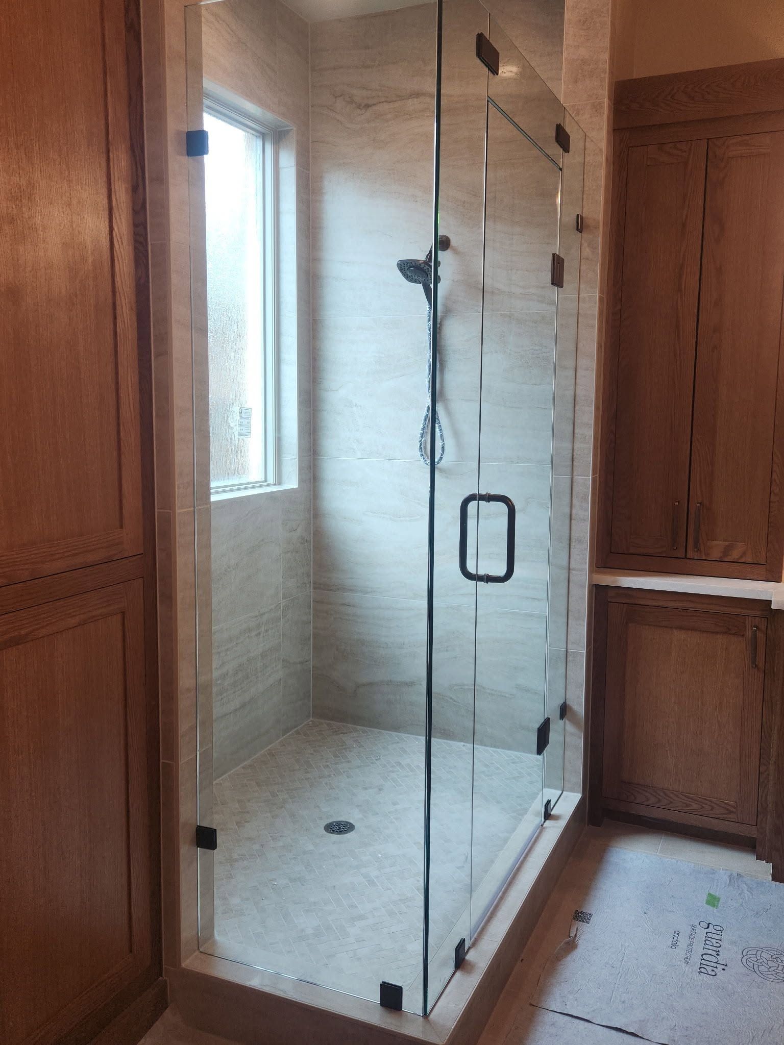 Glass shower enclosure in a beige-tiled bathroom beside a window and wooden vanity.