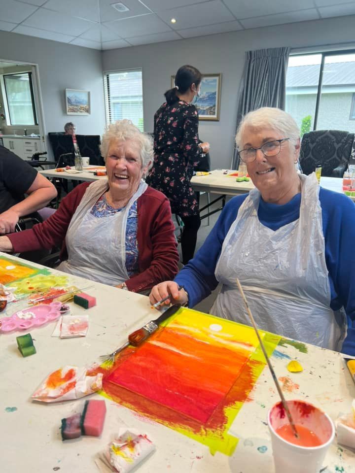 Two older women are sitting at a table painting a picture.