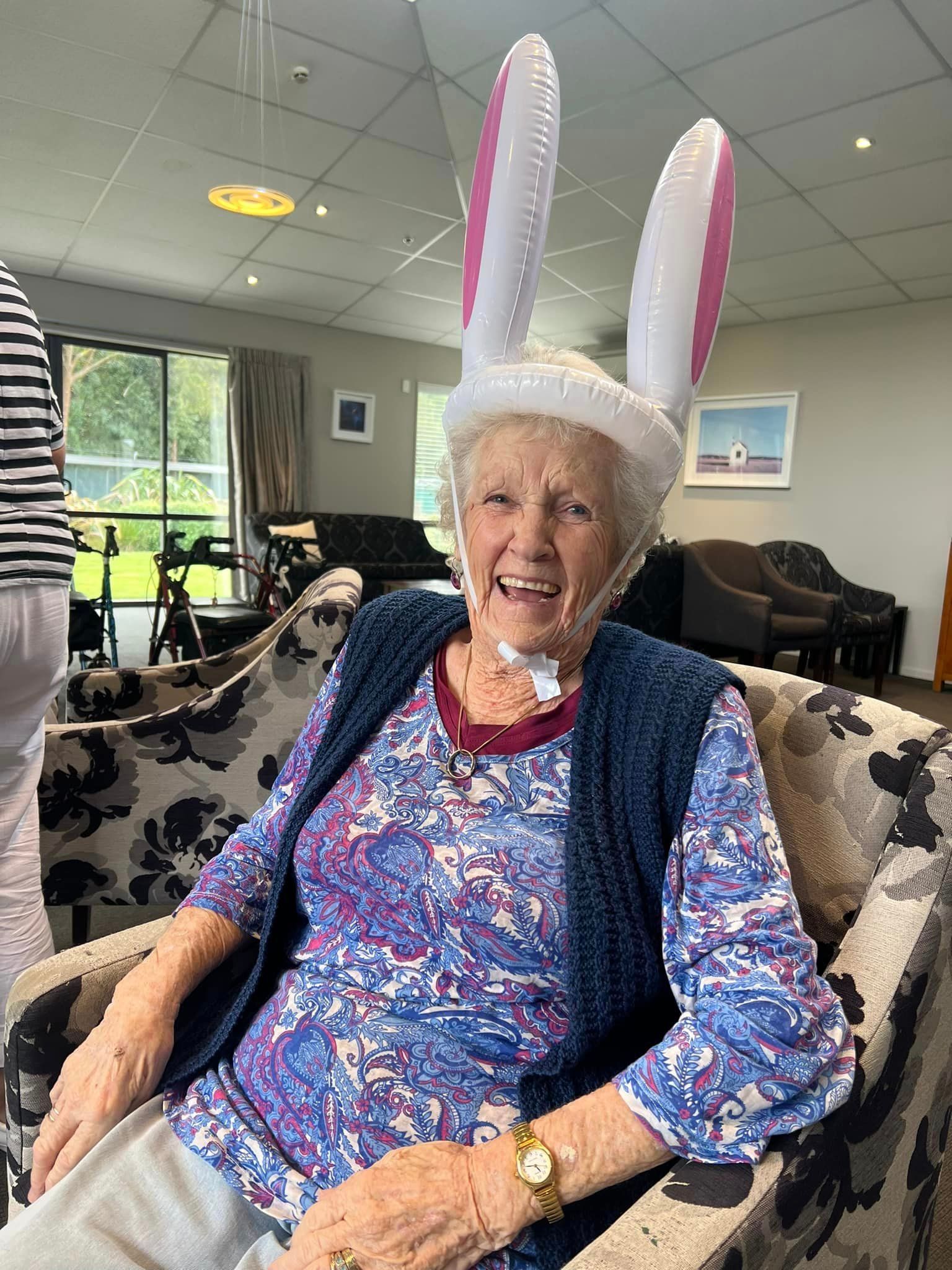 An elderly woman wearing bunny ears is sitting in a chair.