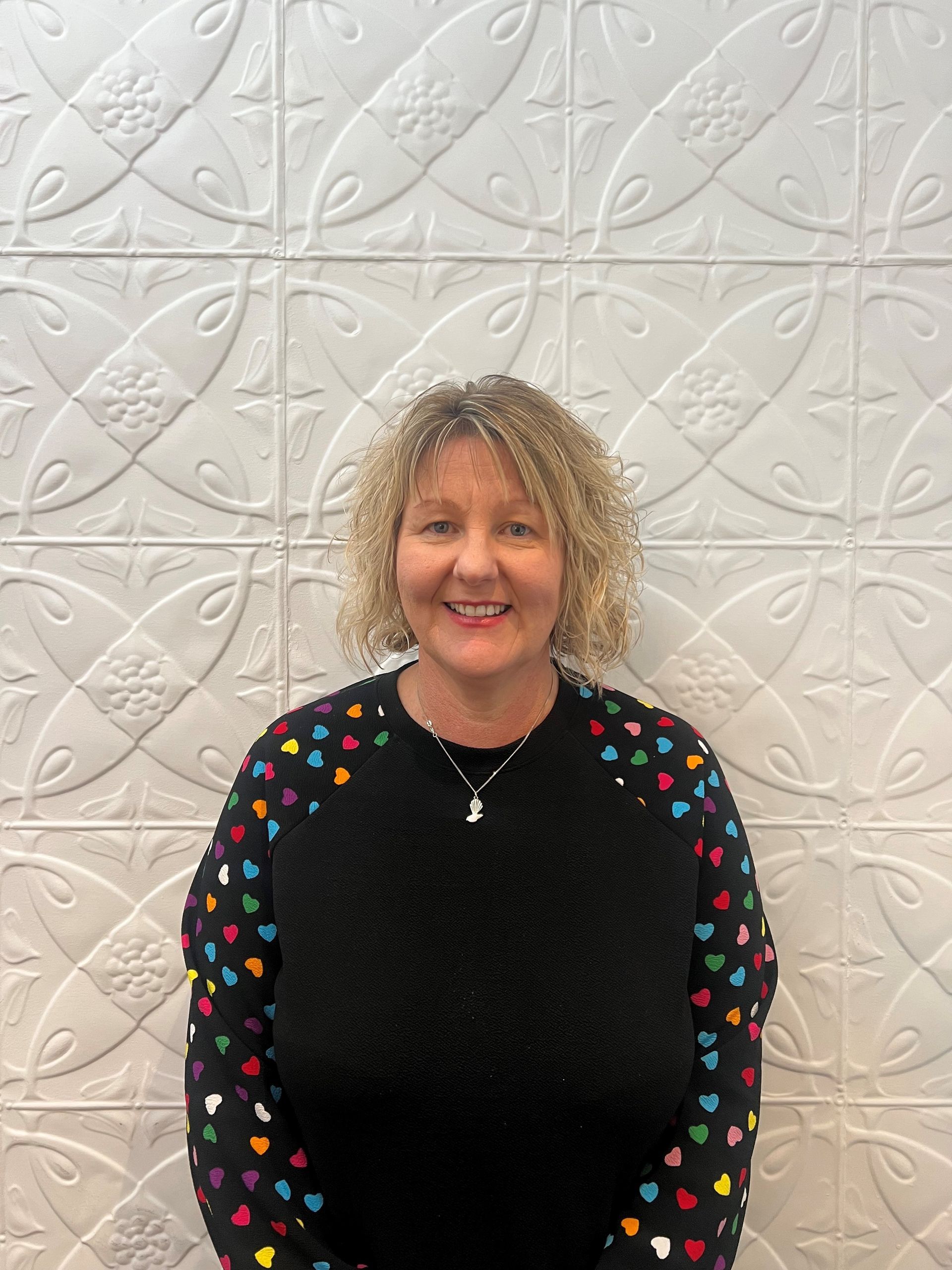 A woman wearing a black sweater with hearts on the sleeves is standing in front of a white wall.
