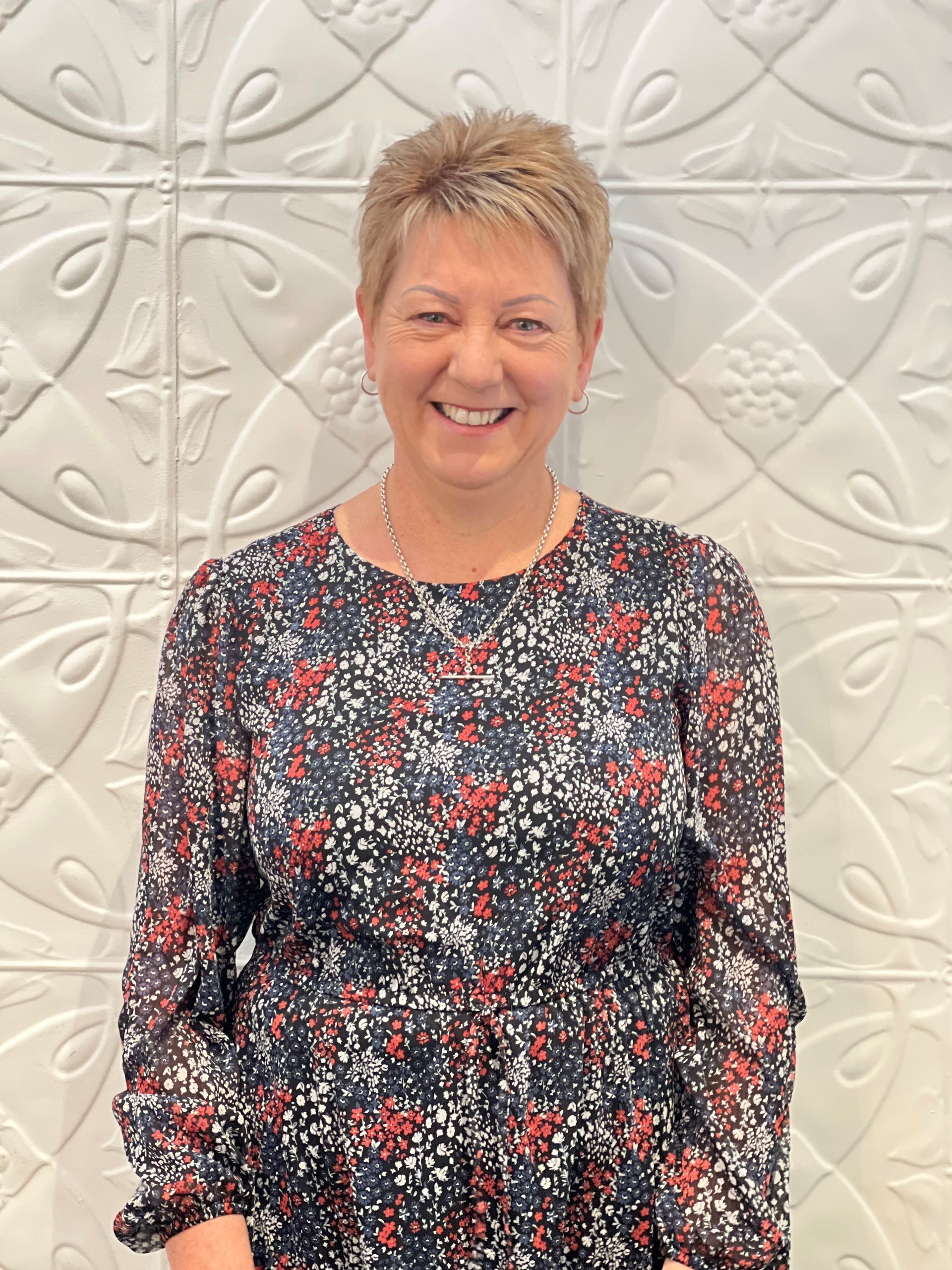 A woman in a floral dress is smiling in front of a white wall.