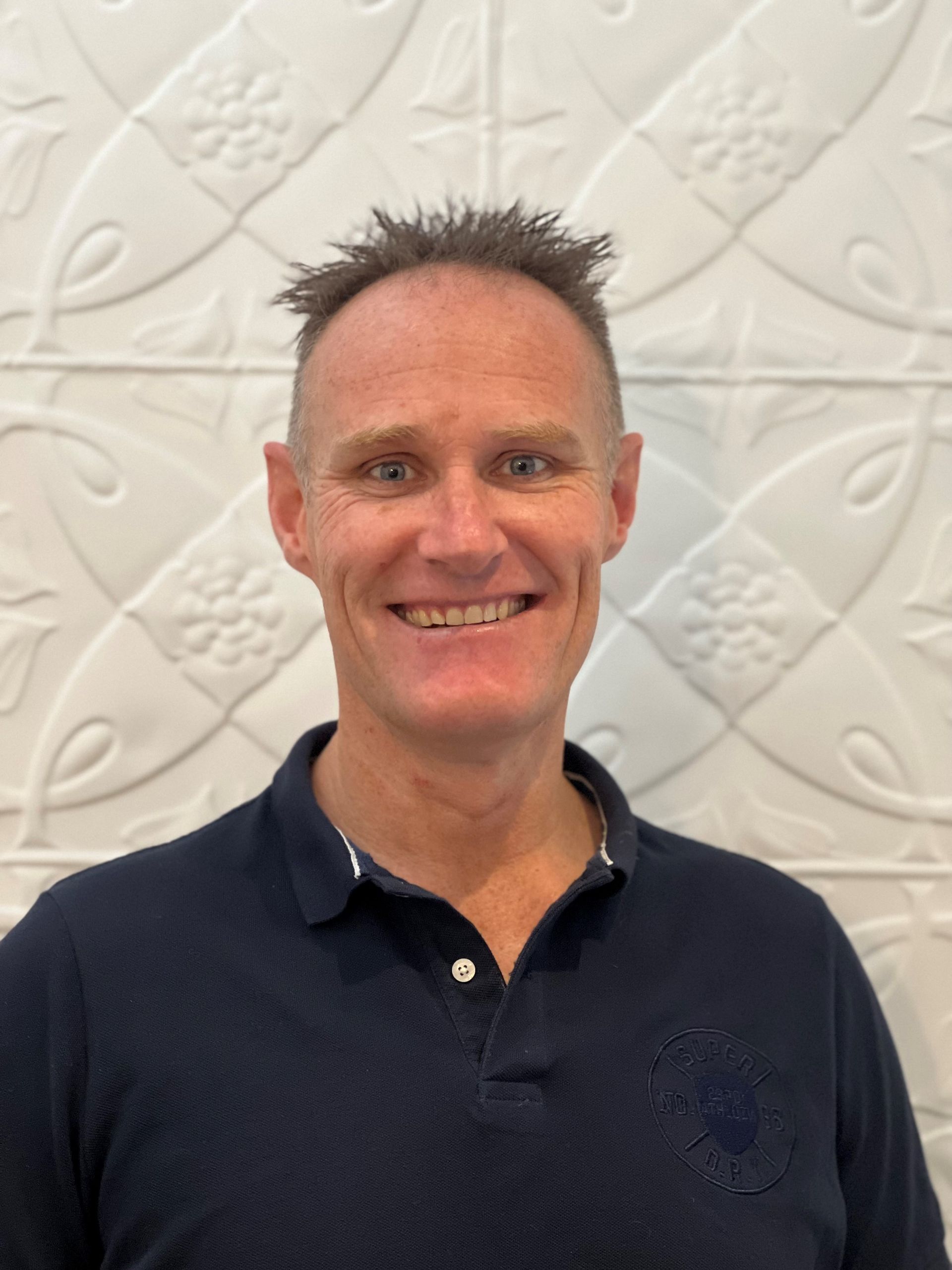A man in a blue polo shirt is smiling in front of a white wall.