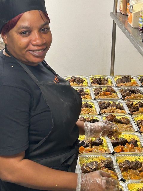 Woman wearing apron and gloves smiling as she prepares meals in aluminum foil containers.