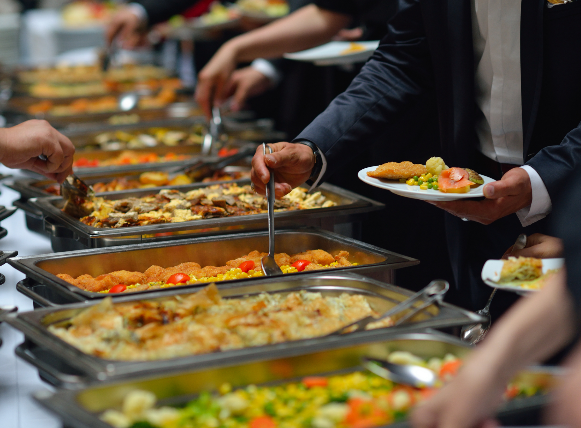 People at a buffet, selecting food with serving spoons. Various dishes in metal trays.