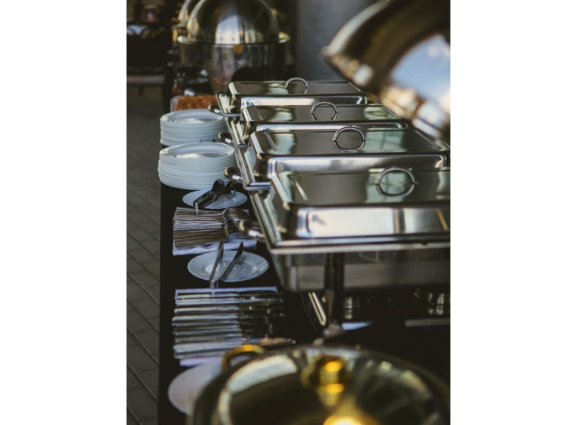Catering buffet table with stainless steel food warmers, white plates, and serving utensils, set outdoors.