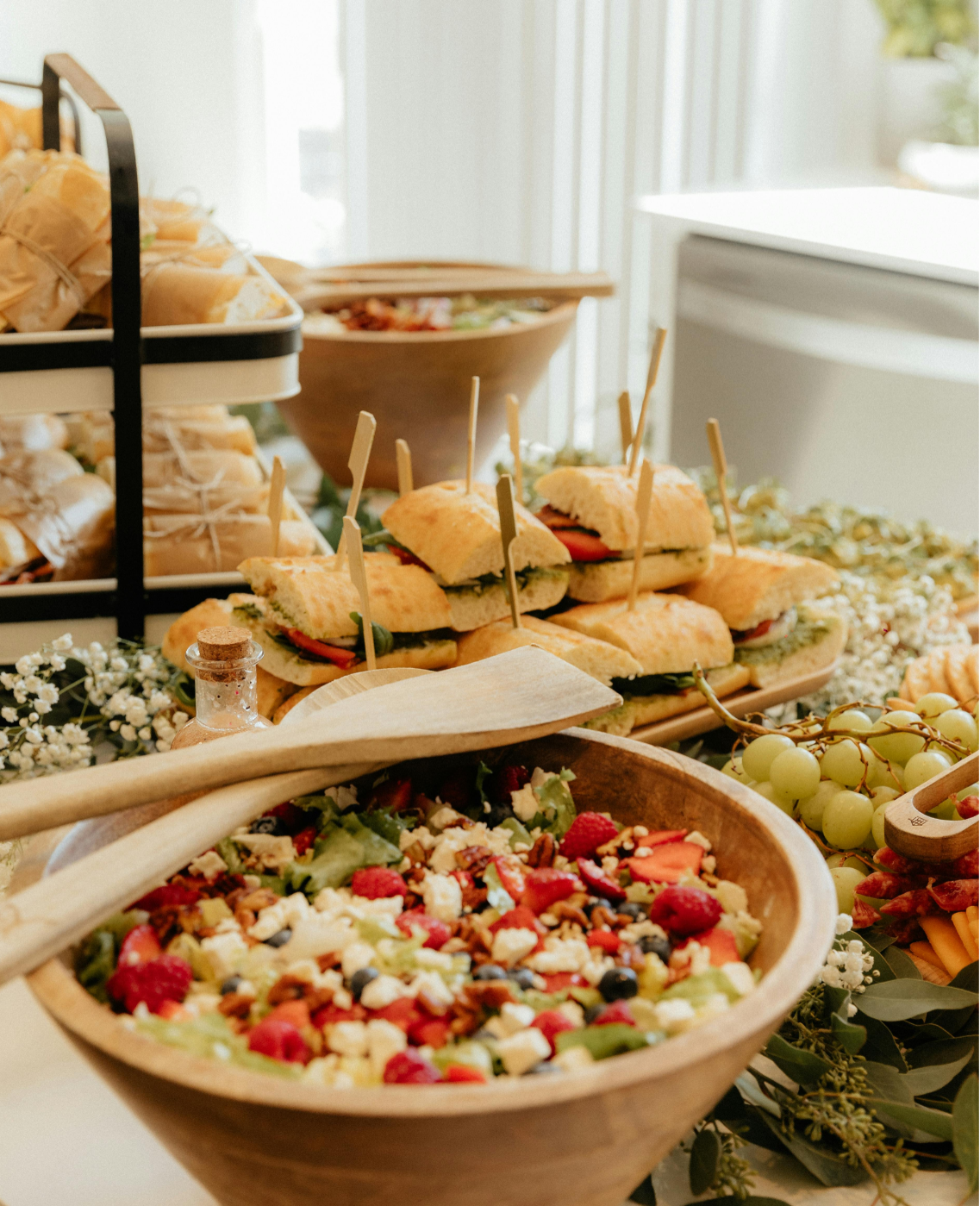 Buffet table with sandwiches, salad, and bread in wooden bowls and a tiered tray.