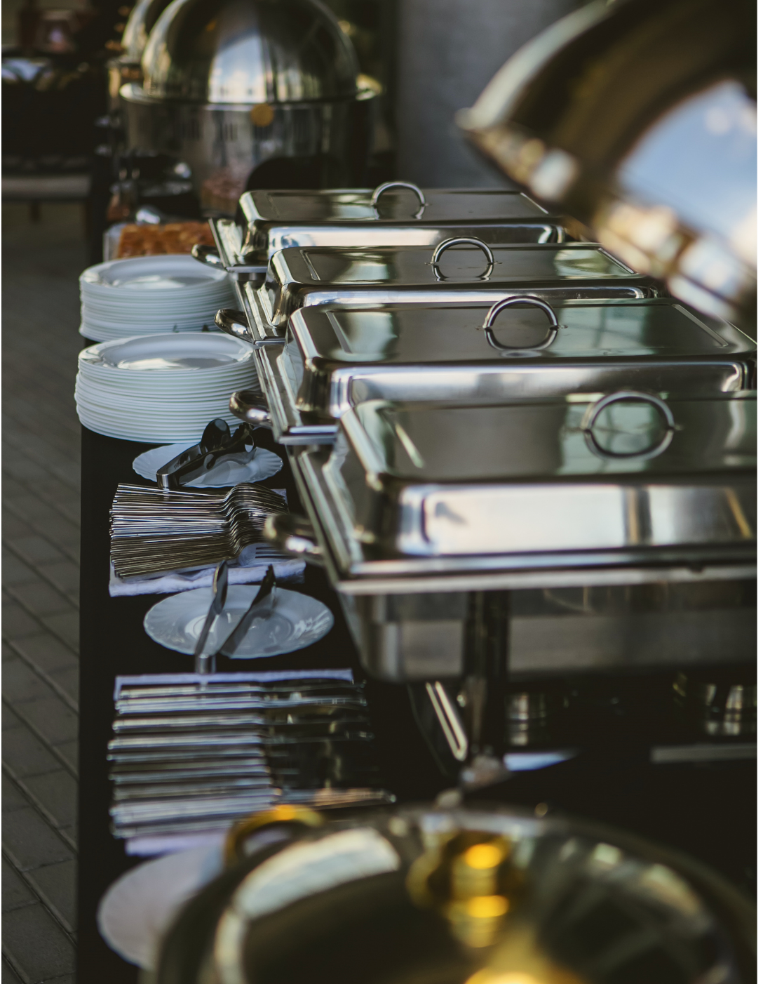 Buffet table with stainless steel chafing dishes and stacks of white plates, setting outdoors.