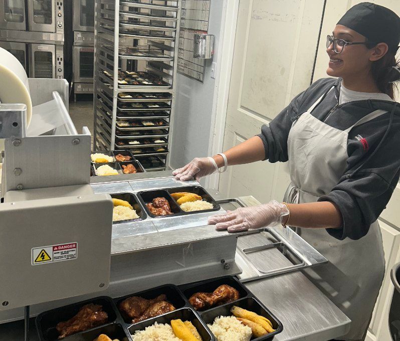 Person sealing prepared food trays with a machine in a commercial kitchen.