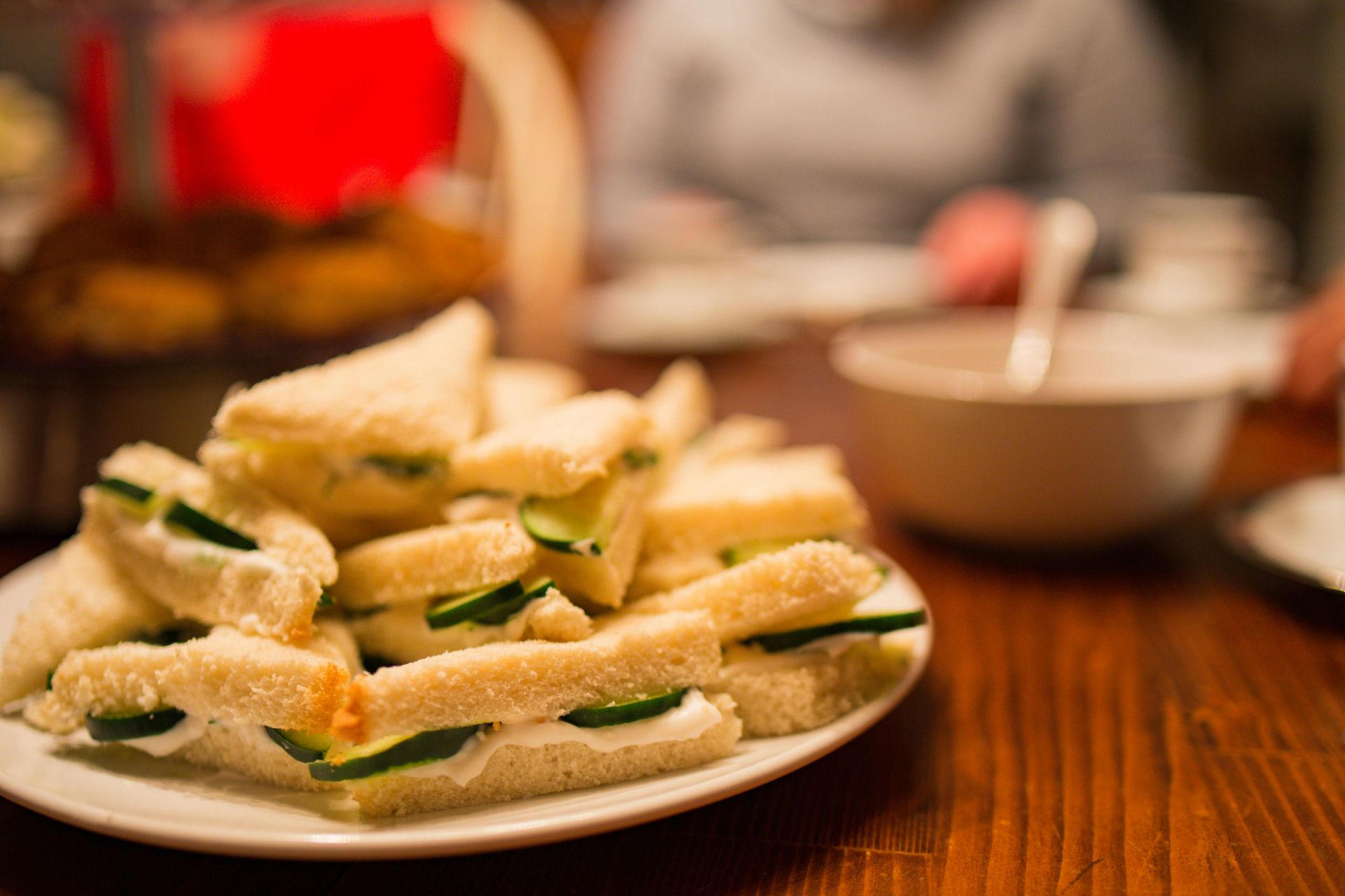 Plate of cucumber sandwiches on a wooden table, with people and food blurred in the background.