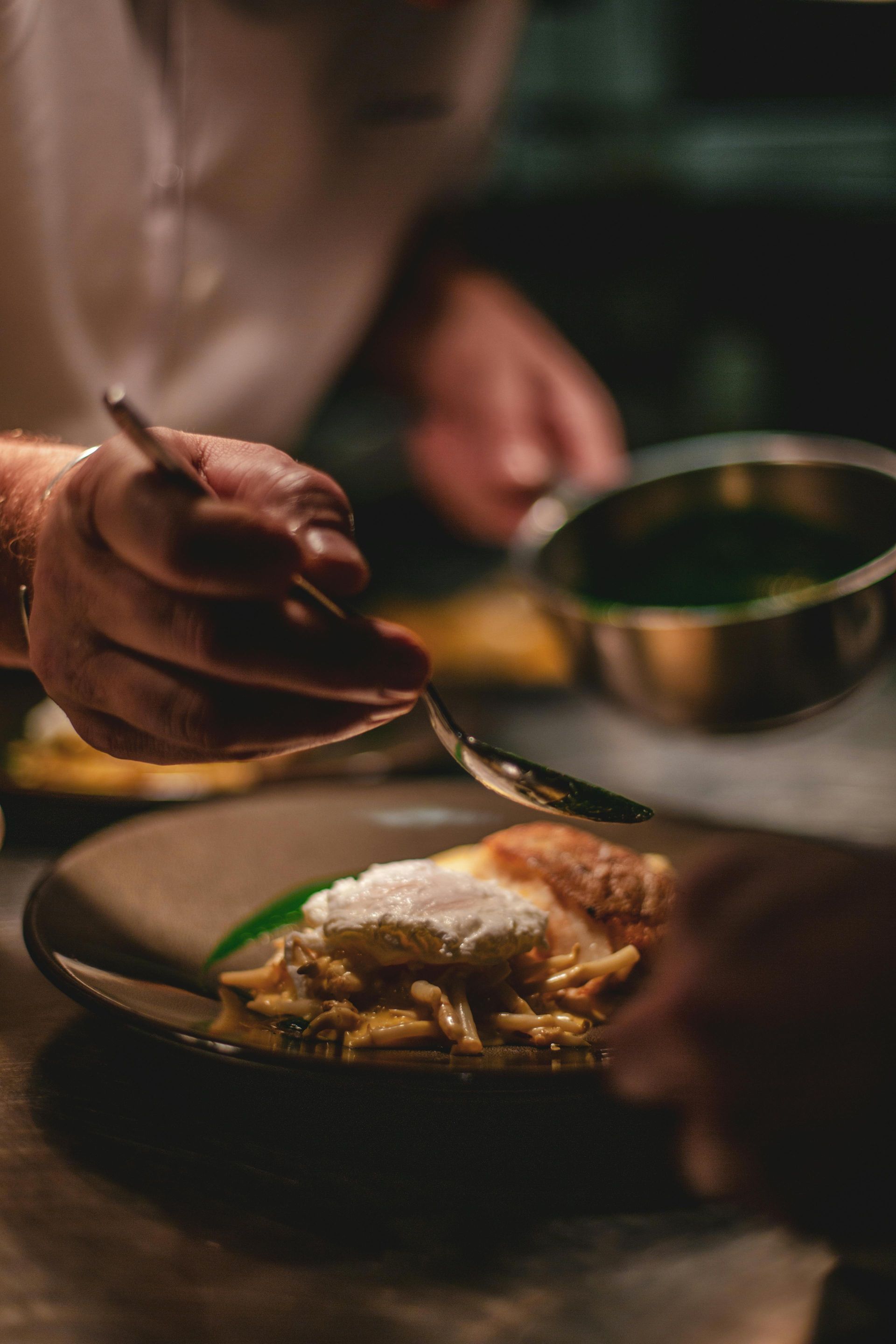 Chef plating a dish with a spoon; cream and sauce on food in a dark kitchen.
