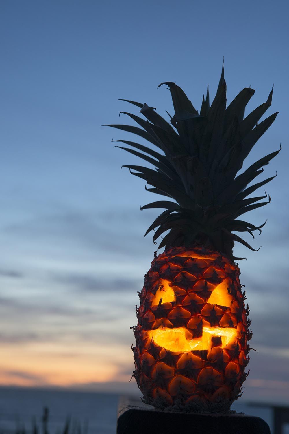 Jack-o'-lantern carved from a pineapple with glowing eyes and mouth, set against a dusky beach sunset.
