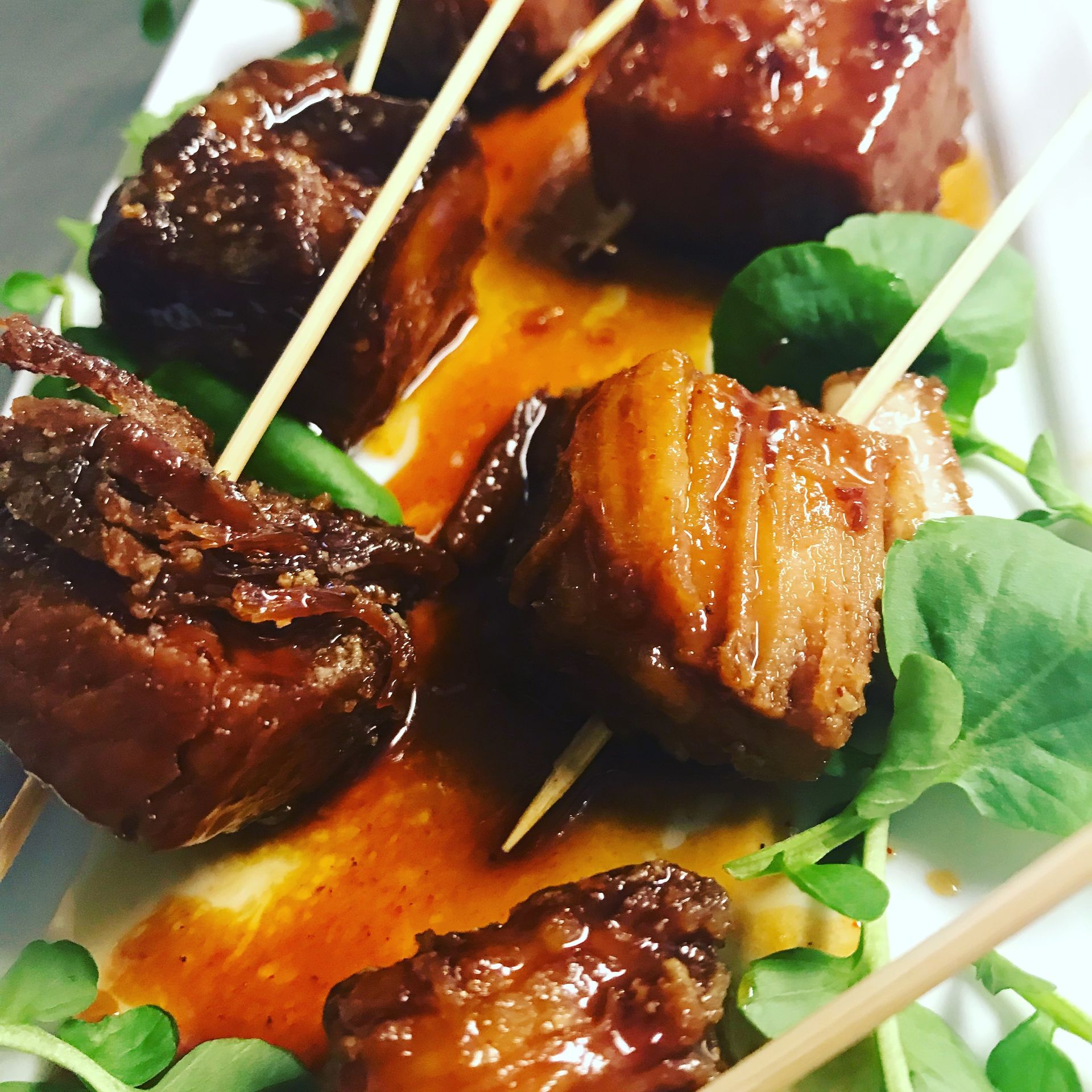 Skewered glazed pork belly bites on a white plate, garnished with greens and sauce.