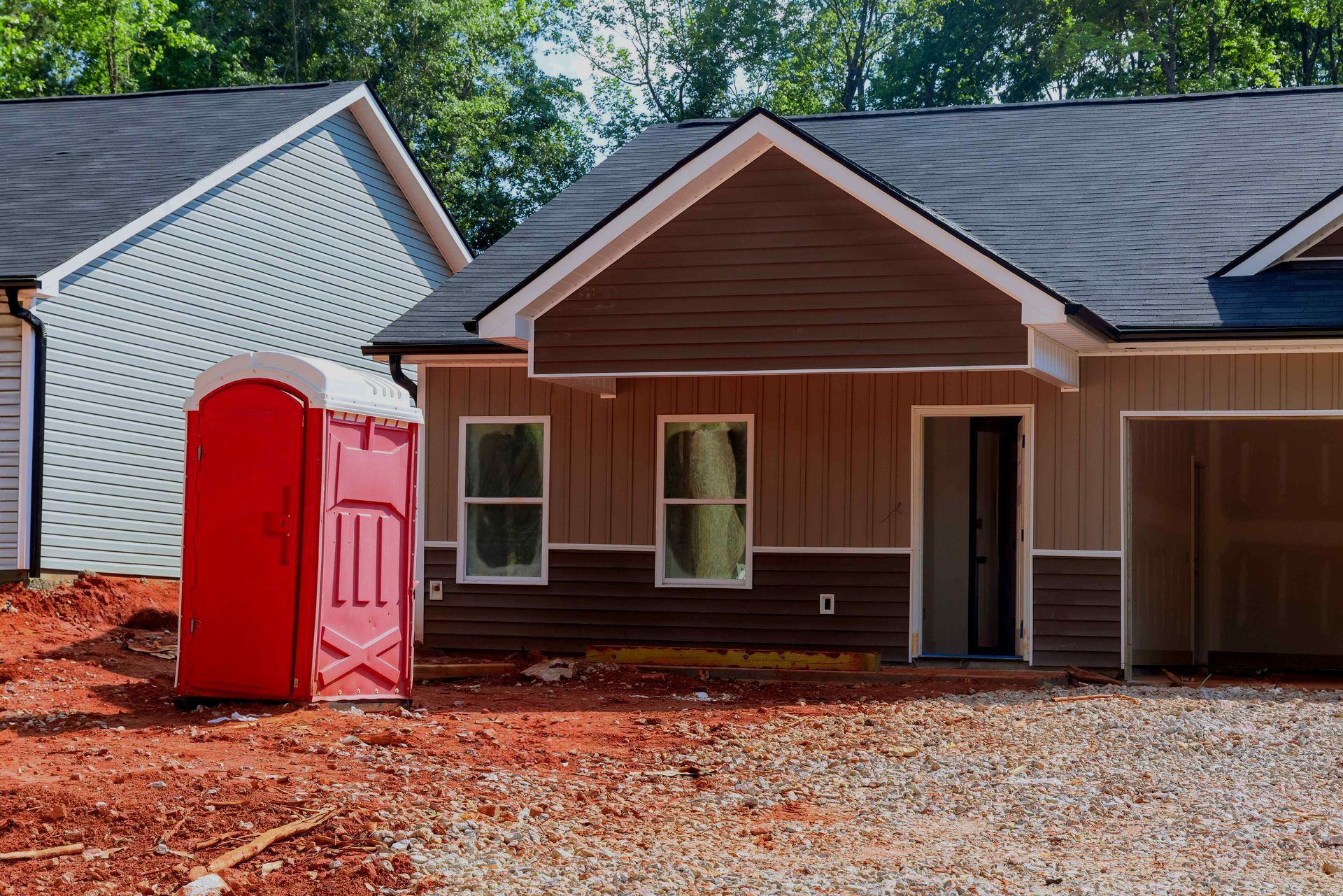 A red portable toilet is parked in front of a house under construction.