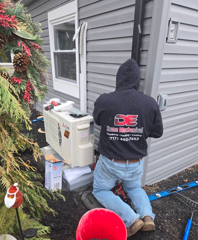 A person in a hooded sweatshirt kneels outside, working on a unit near a building.