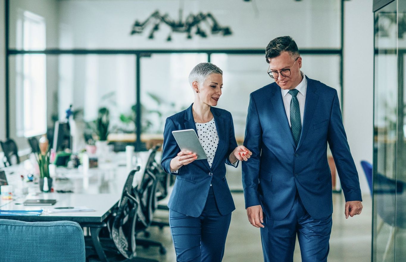 A man and a woman are walking through an office while the woman is holding a tablet.