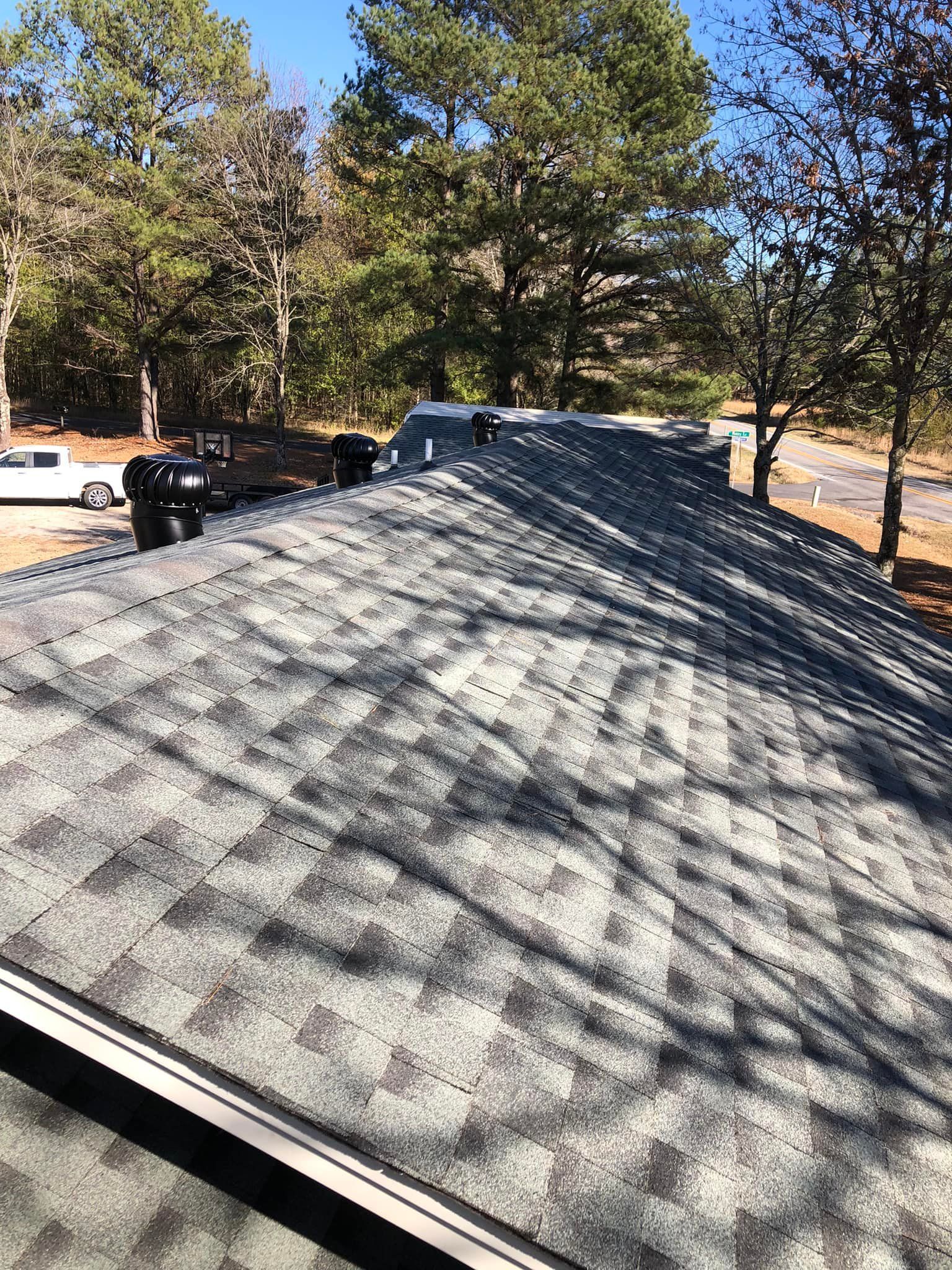 New asphalt shingle roof on a house, under a sunny sky, with trees in the background.