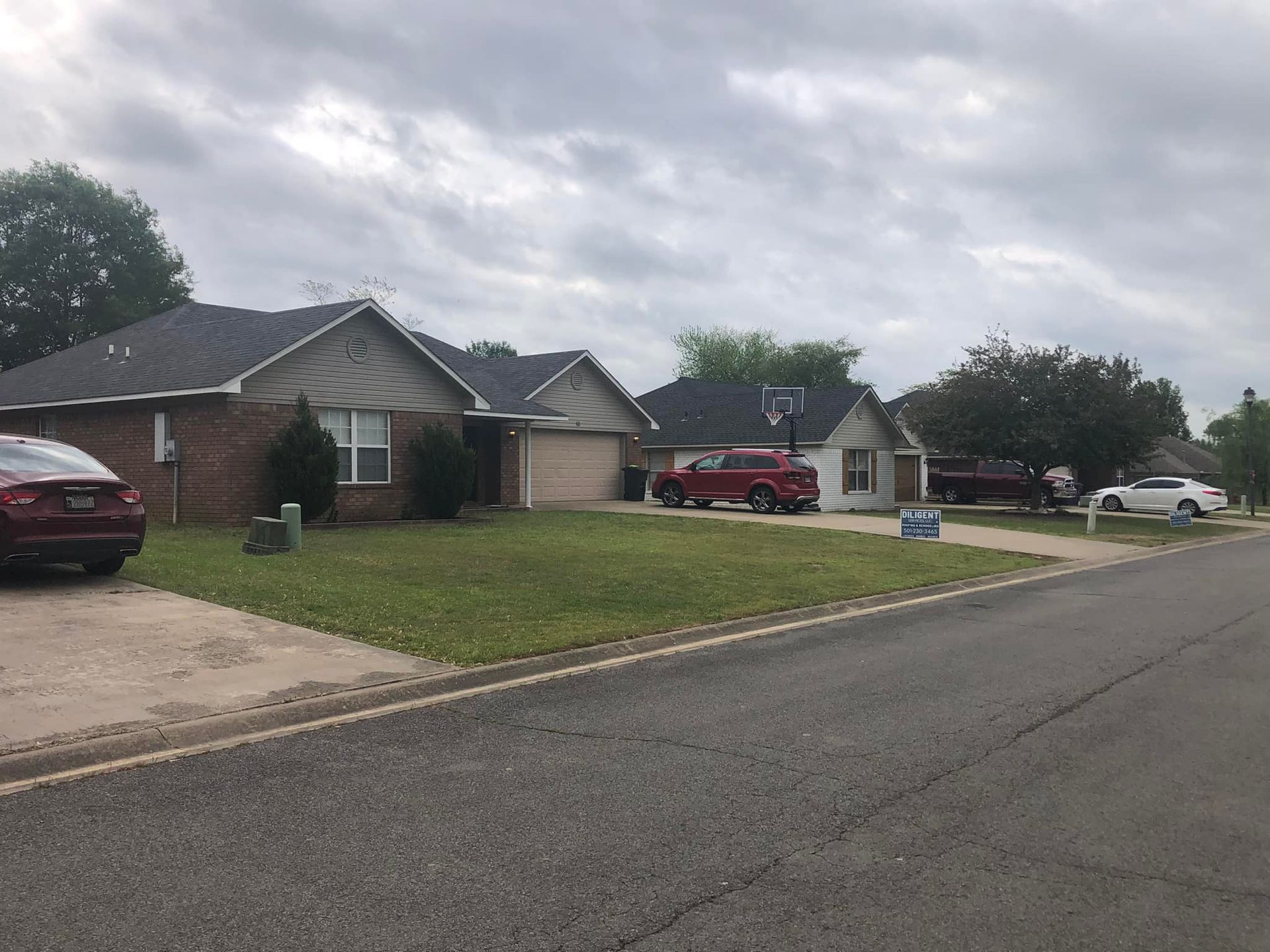 Houses along a suburban street under a cloudy sky. Cars are parked in driveways and on the street.