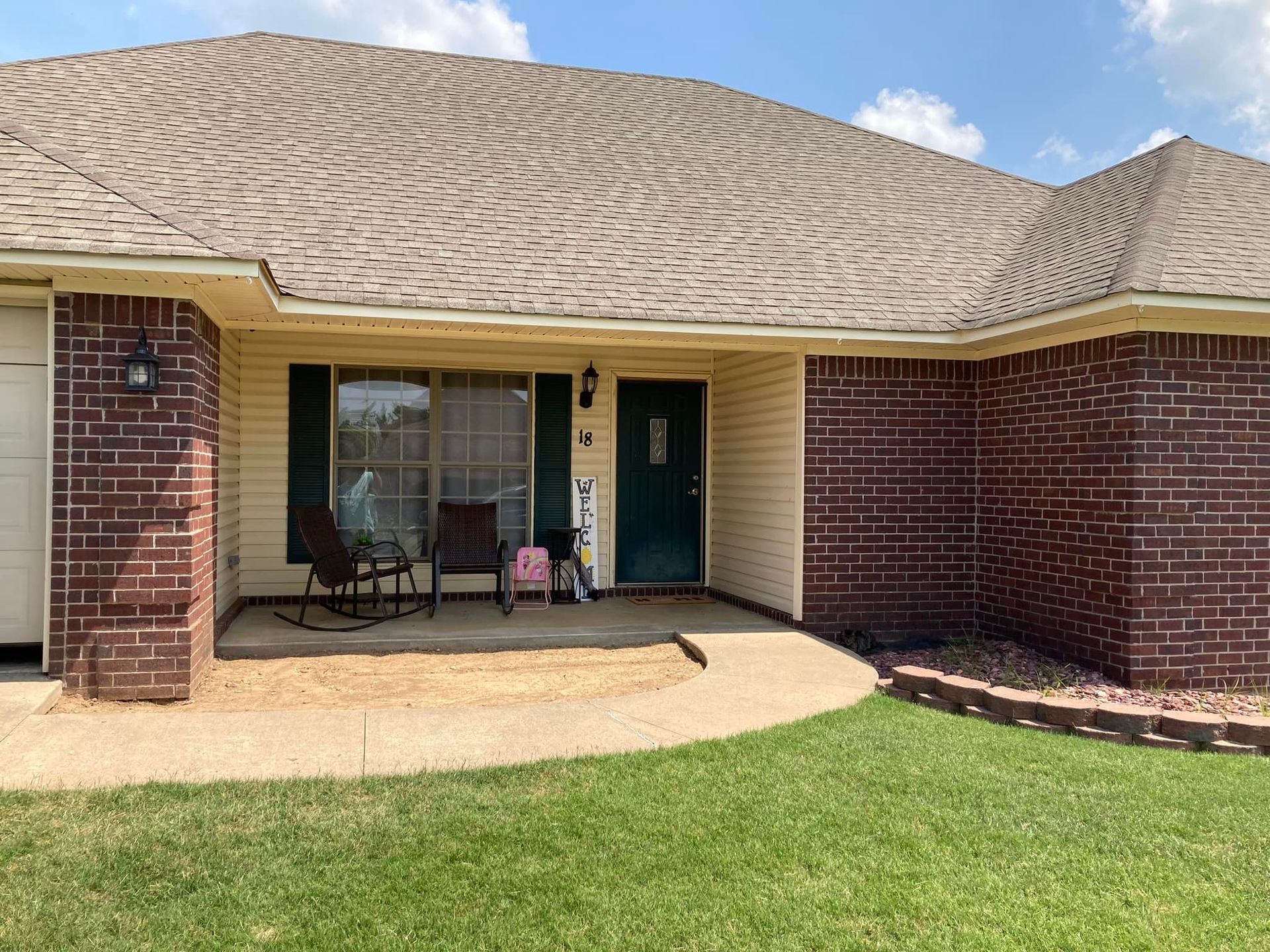 Tan house with brown roof and brick accents, small porch with chairs, green lawn.