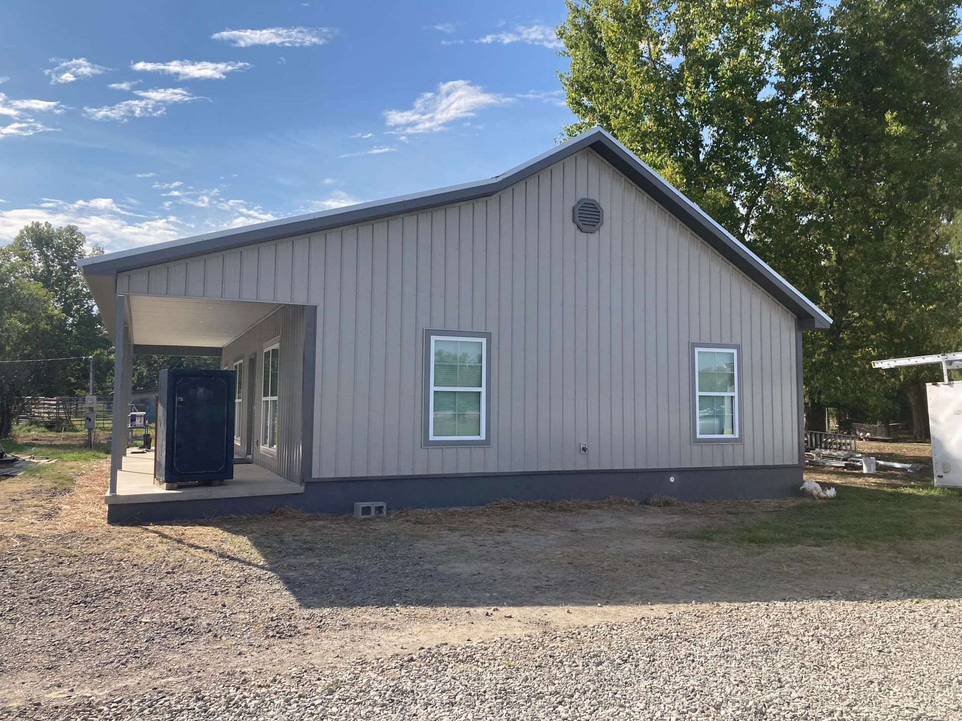 Gray metal-clad house with a porch and two windows, set on a gravel lot under a partly cloudy sky.
