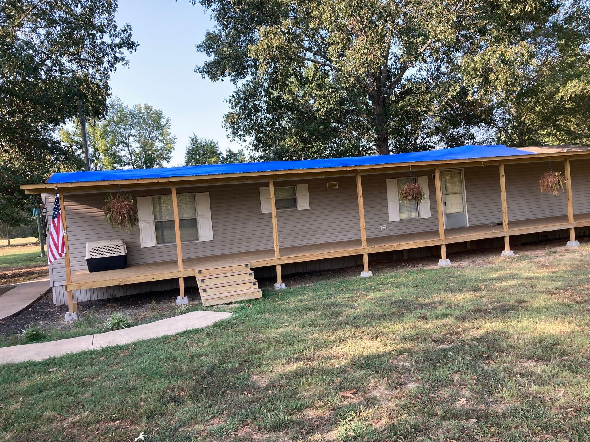 Mobile home with a blue tarp roof and sloped wooden porch; American flag.