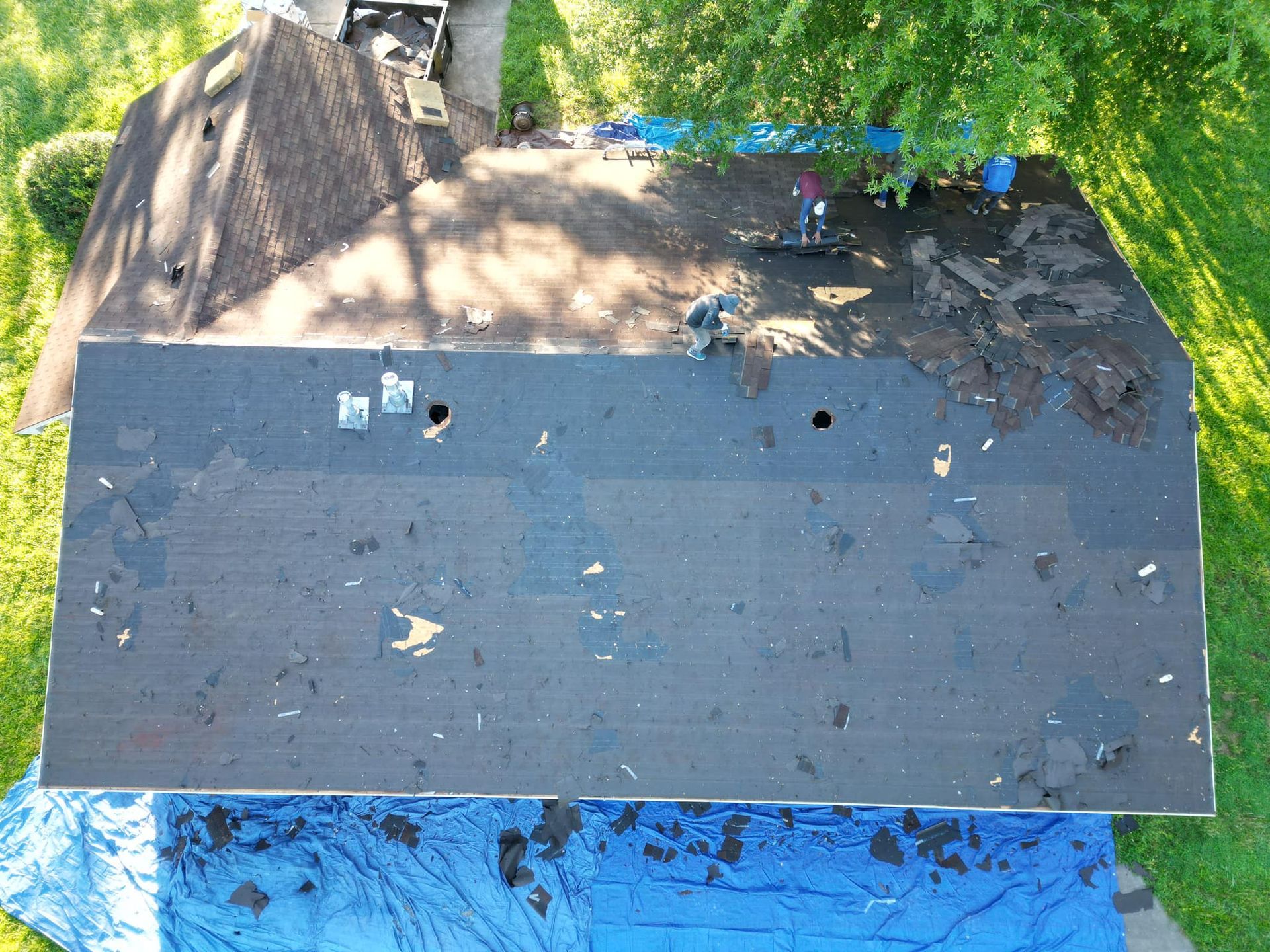 Overhead view of a house roof partially stripped during repair, with a worker and tarps visible.