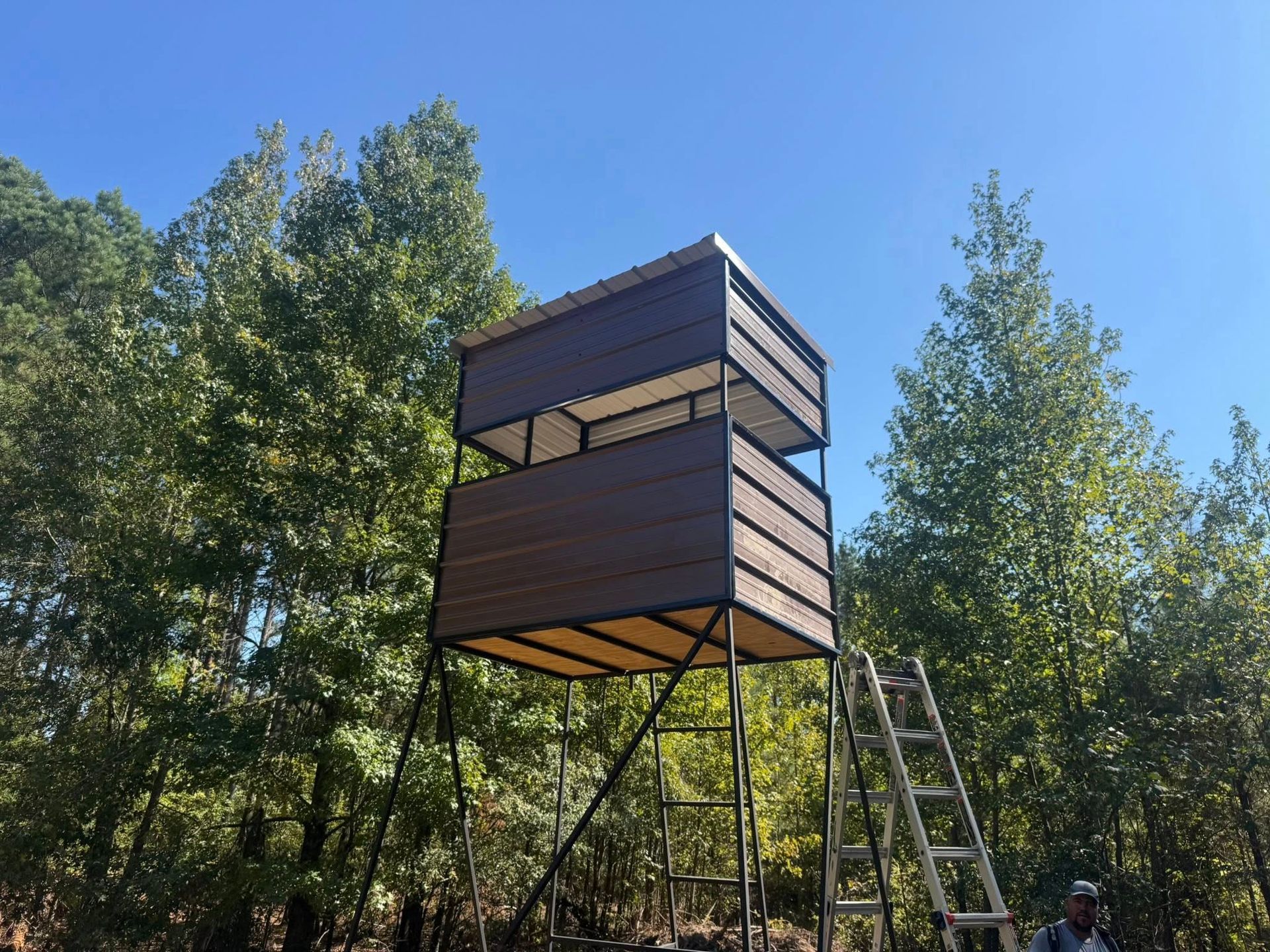 A tall, brown hunting blind with ladder in a wooded area, under a blue sky.