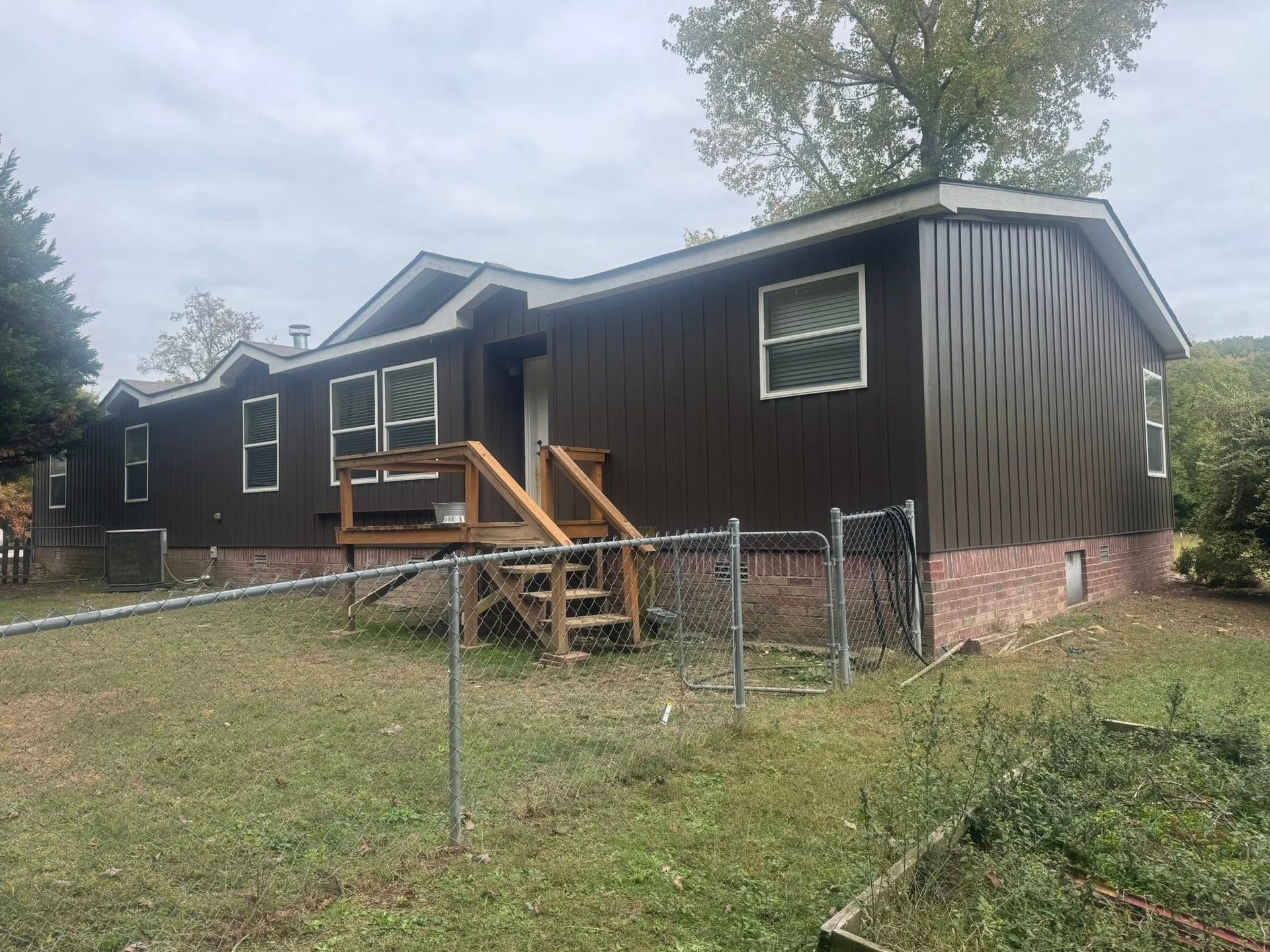 Brown house with wooden steps, surrounded by a chain-link fence on a cloudy day.