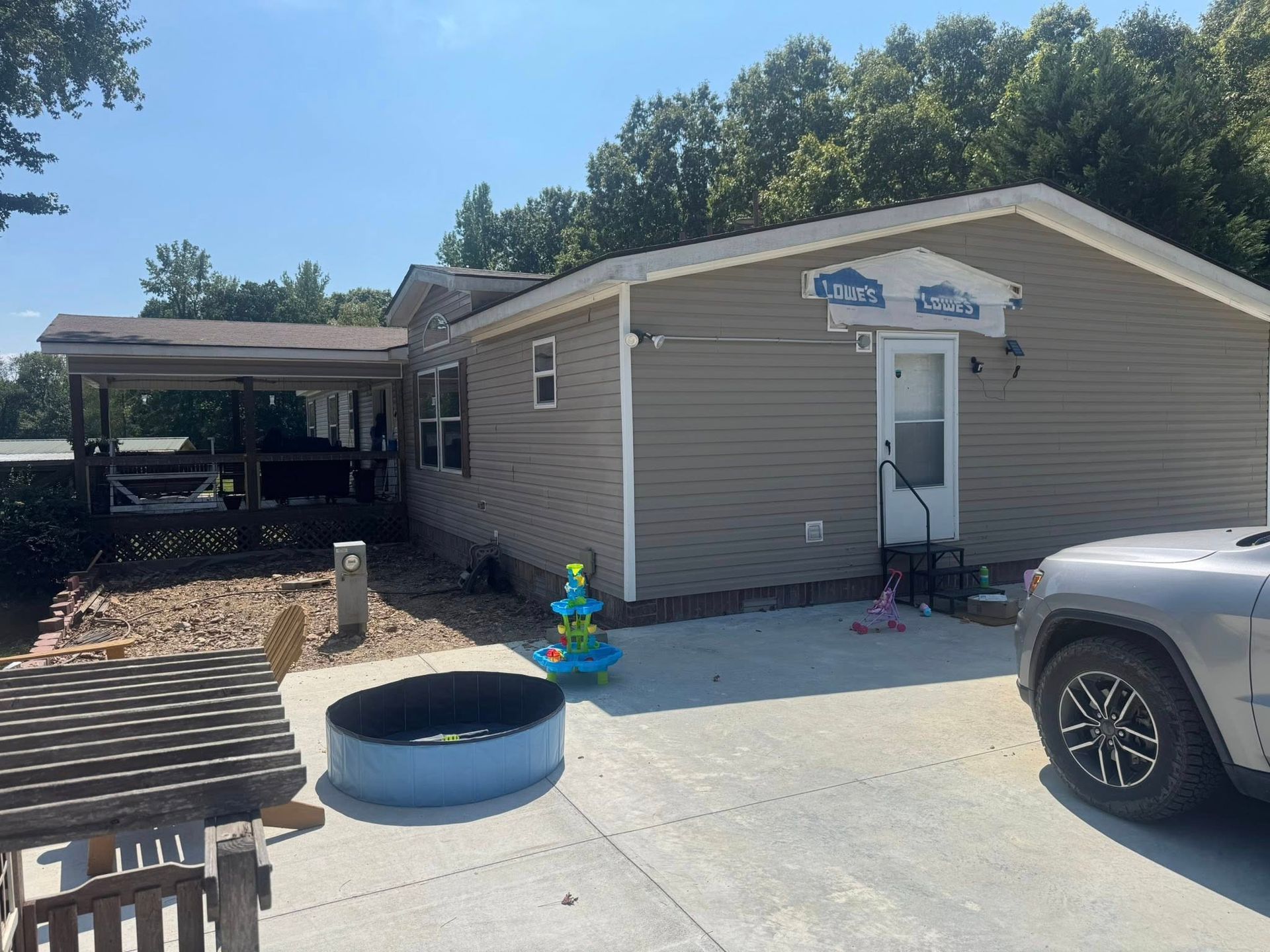 Exterior view of a beige house with a carport and driveway; a kiddie pool and Jeep are visible in the yard.