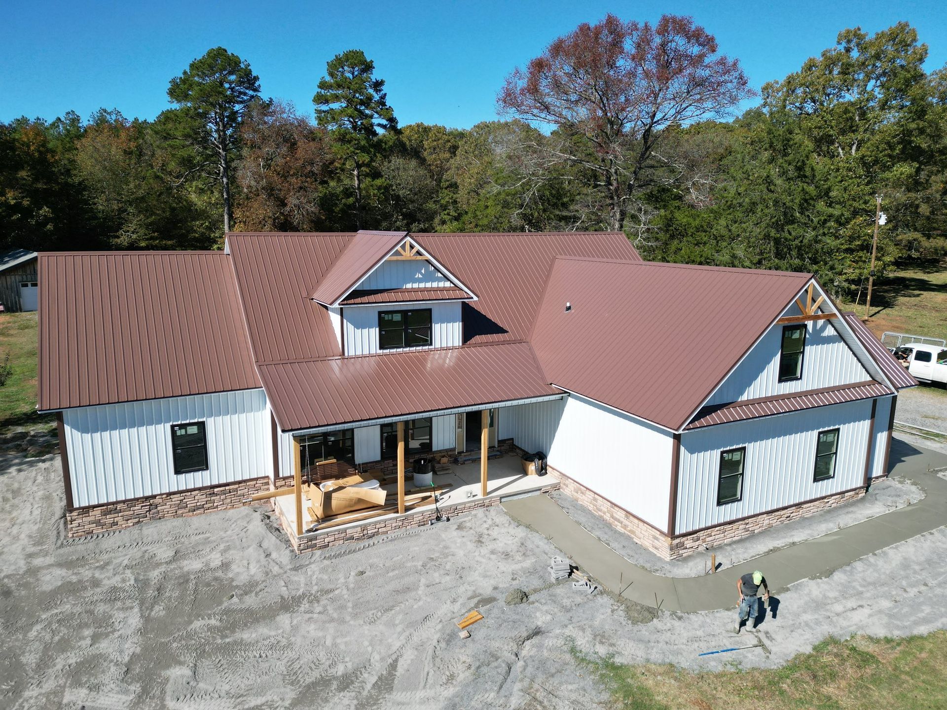 Brown-roofed, white farmhouse under construction with porch and two workers; trees in background.
