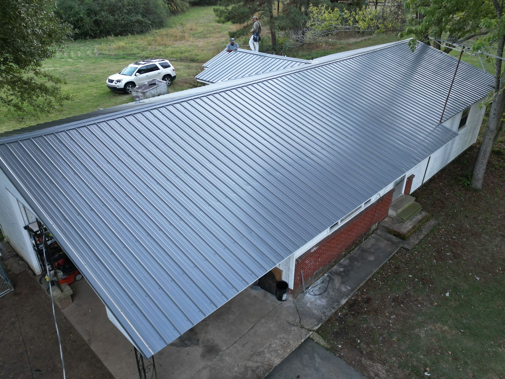 Gray metal roof on a white and brick house. A car is parked on the grass beside it.