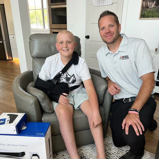 A boy with a broken arm is sitting in a chair next to a man wearing an amsl shirt