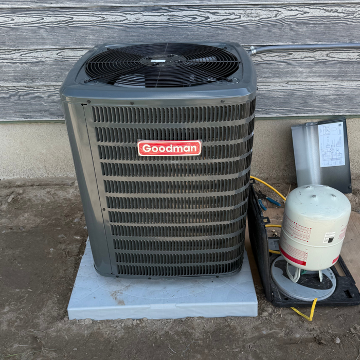 Gray Goodman air conditioner unit with attached equipment, sitting outside on a concrete pad.
