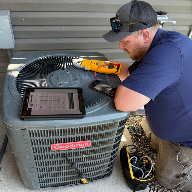HVAC technician inspecting a Goodman AC unit with a multimeter, tablet, and phone outside.